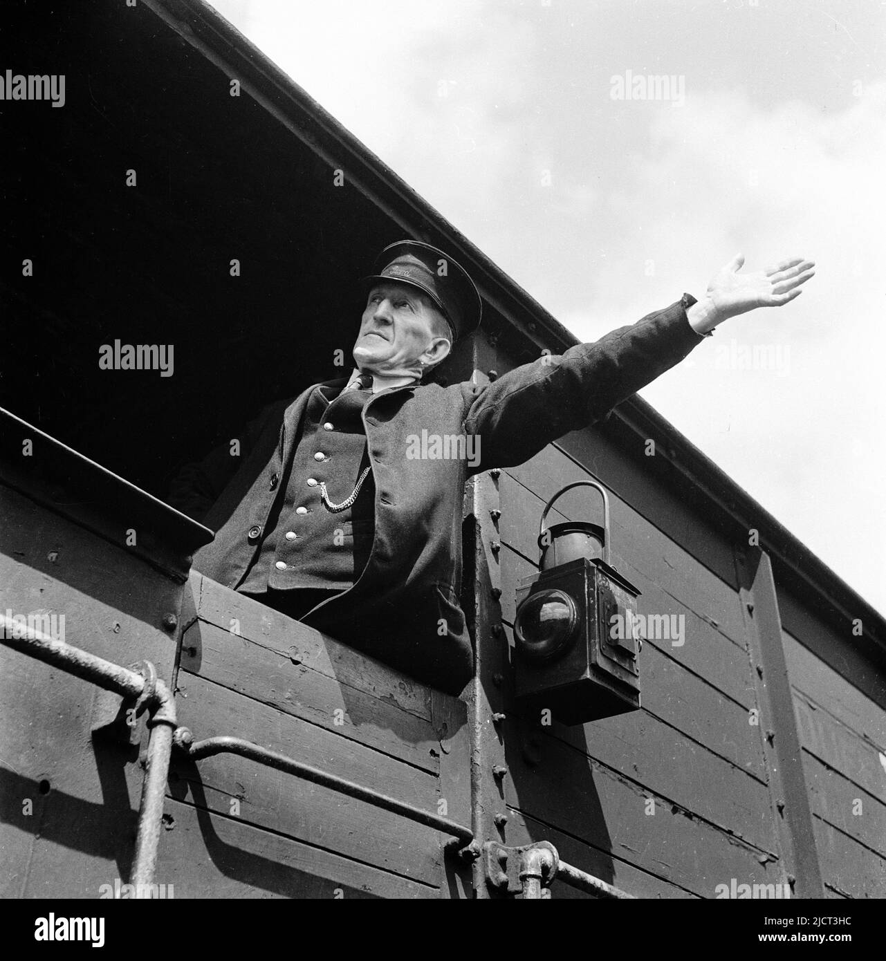 1950s, historisch, ein uniformierte Zugwächter, der in einem hölzernen gerahmten Güterwagen steht und mit einem ausgestrecktem Arm ein Handzeichen gibt, dockt London, England, Großbritannien an. Das Wort Wache ist auf seinen Arbeitshut genäht. An der Außenseite des Wagens hängt eine Bahnleuchte. Stockfoto