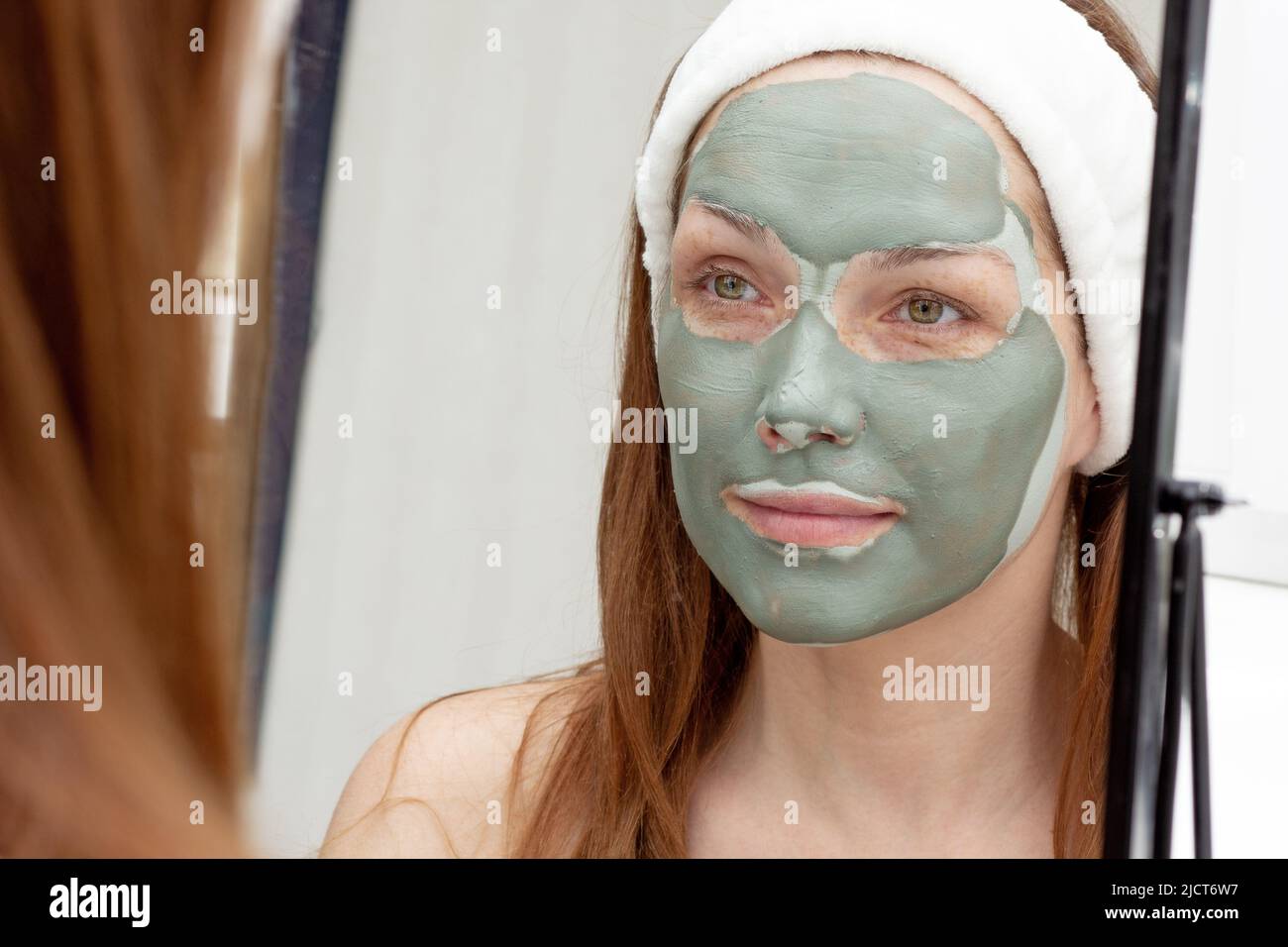 Lächelnde Frau mittleren Alters mit Band auf dem Kopf und Blick auf ihr Gesicht mit blauer Tonmaske im Spiegel Stockfoto