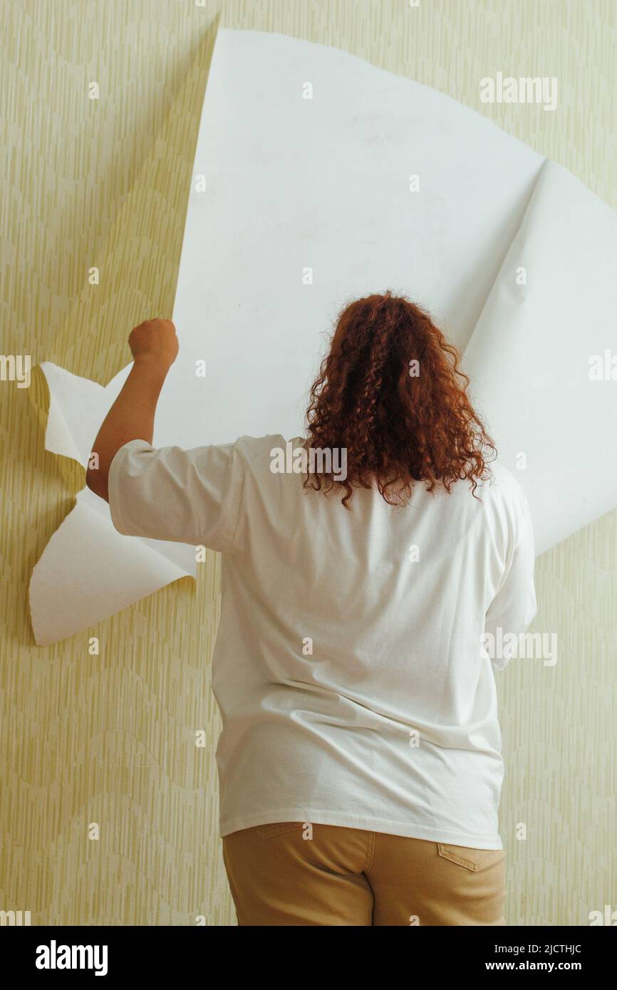 Rückansicht einer fetten Frau mit weißem T-Shirt, beigefarbener Hose, Ständer, der gelbe Tapete von der Wand abreißt. Vertikal. Stockfoto