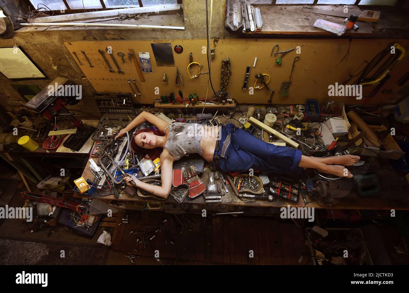 Eine junge, schöne Frau findet während ihrer Arbeitszeit Zeit zum Schlafen. Sie legt sich auf den Arbeitstisch, der mit Werkzeugen und anderen elektronischen Geräten gefüllt ist. Stockfoto