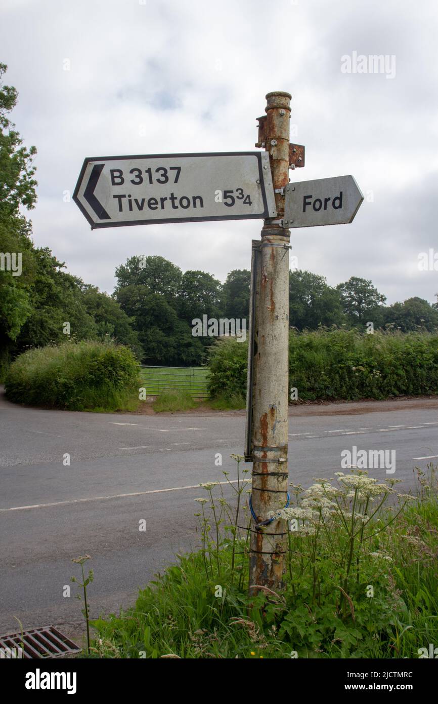 Weißes Straßenschild nach Tiverton und Ford mit einer Straße und Hecken im Hintergrund Stockfoto