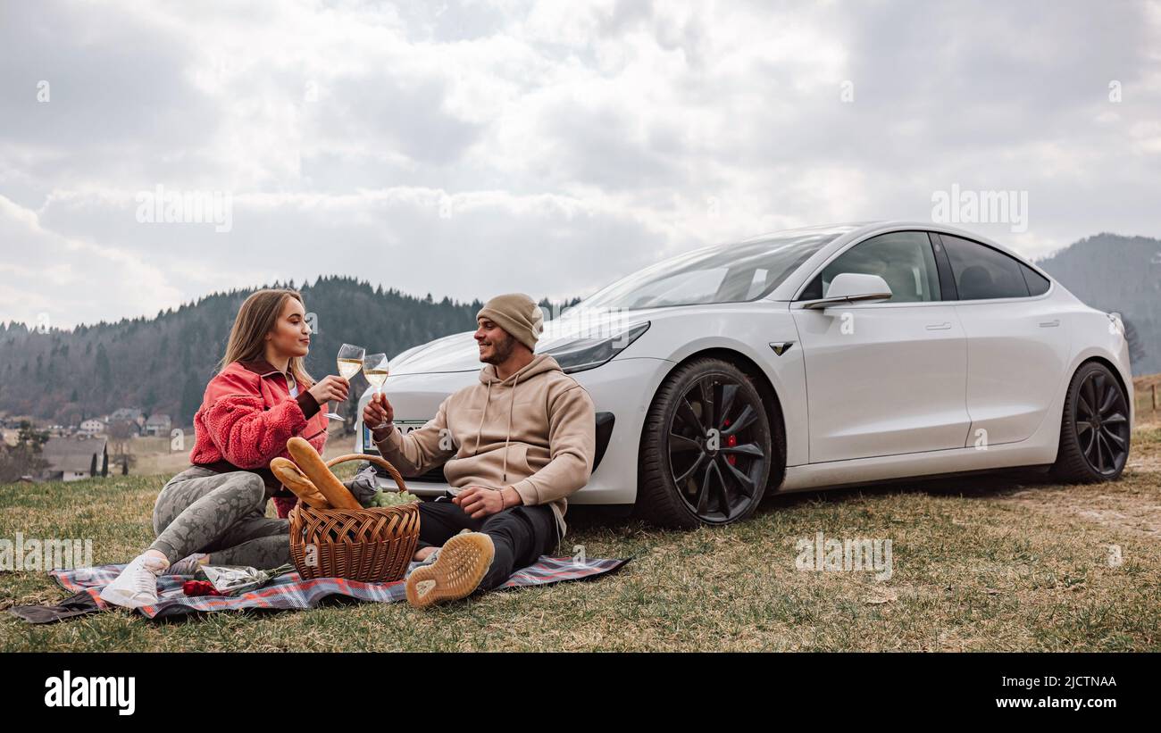Lächelndes junges, modernes Paar, das eine Reise in die Natur macht, Wein und Trauben genießt, während es entspannt neben ihrem neuen exklusiven Elektroauto sitzt Stockfoto