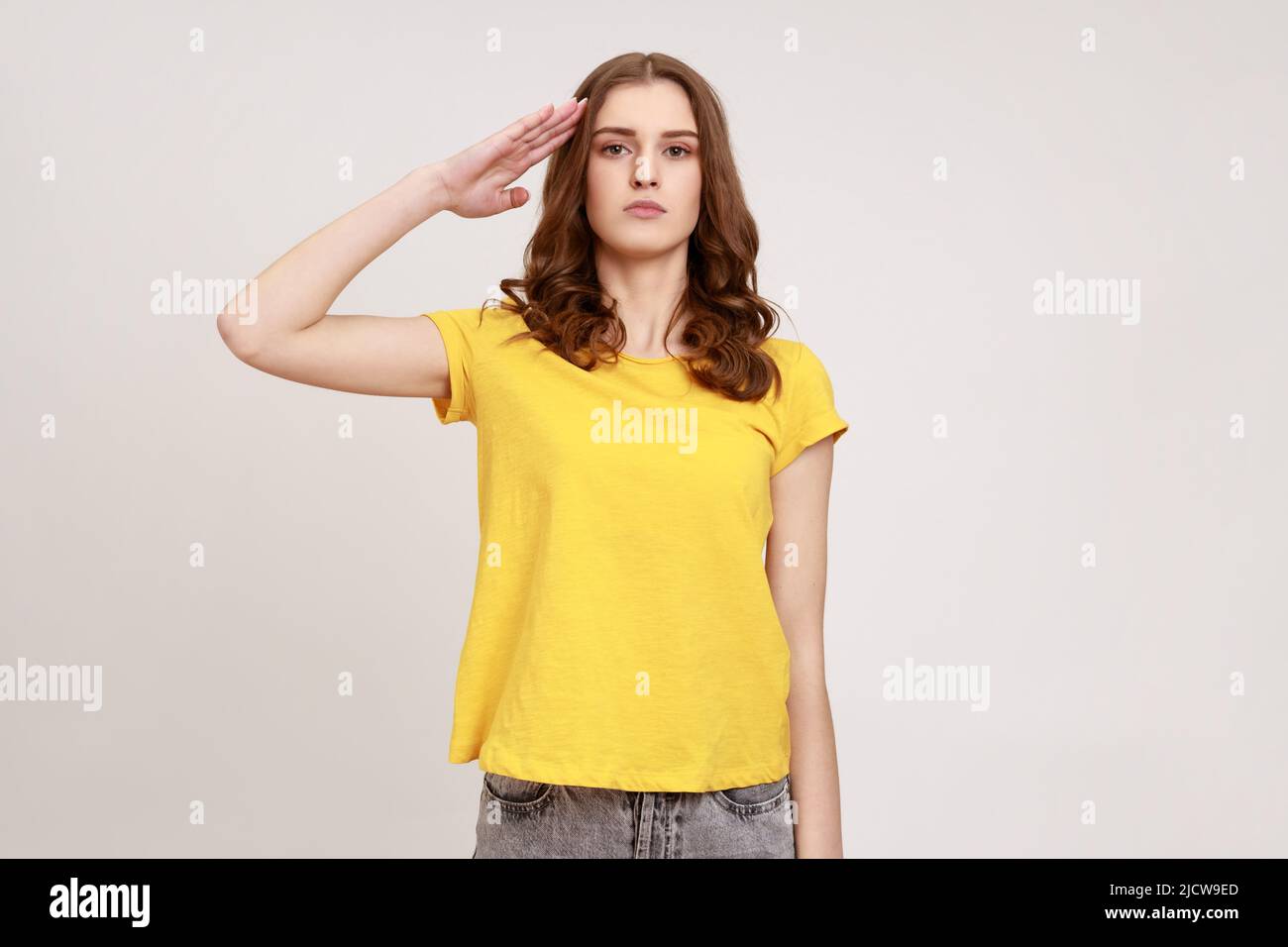 Ja, Sir. Junge patriotische Mädchen in beiläufigen gelben T-Shirt Hand in der Nähe des Kopfes halten, grüßend, Ehre und Ruhm, ernsthaft in die Kamera schauen. Innenaufnahme des Studios isoliert auf grauem Hintergrund. Stockfoto