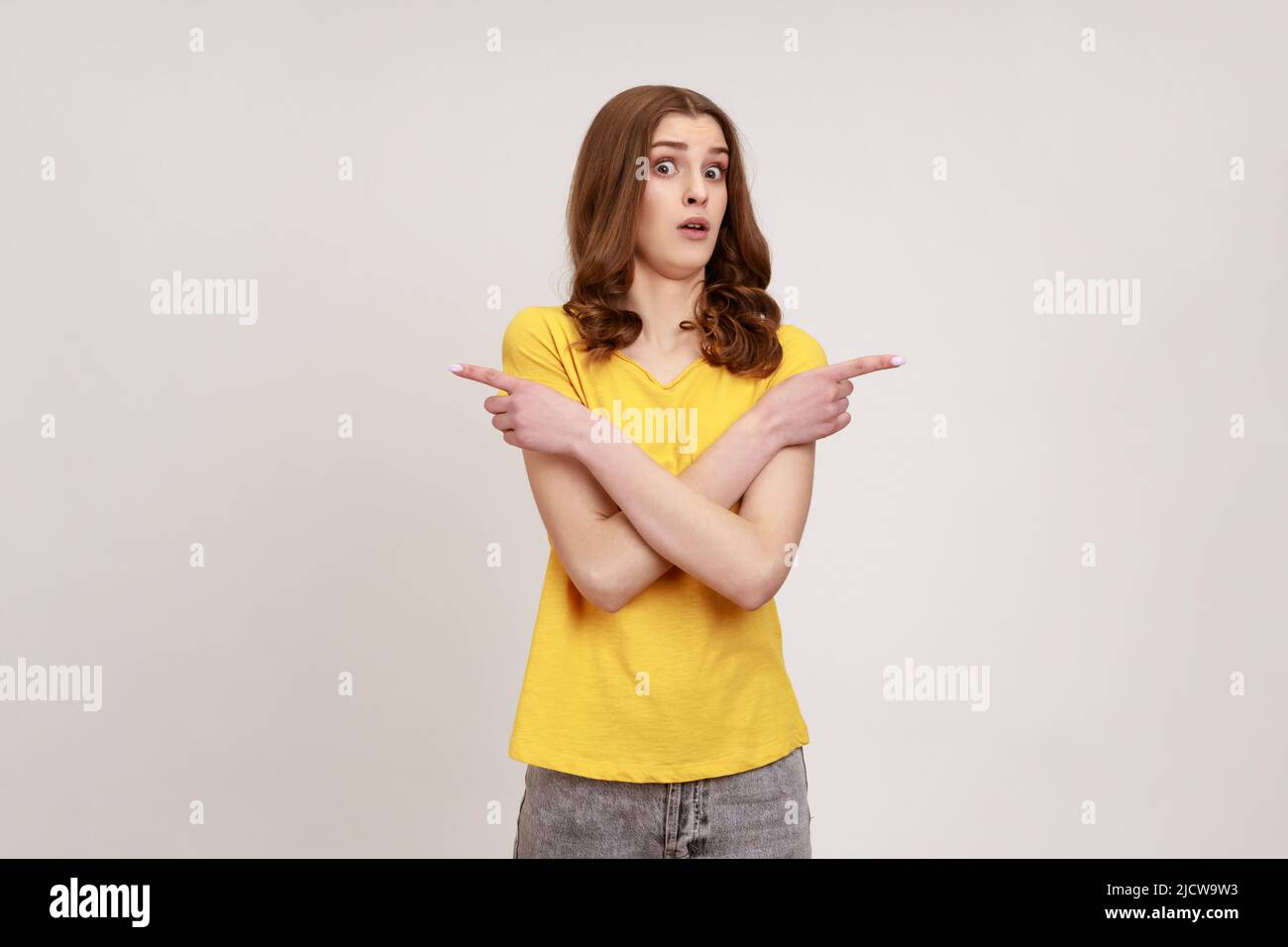 Porträt von verwirrt verwirrt Teenager Mädchen tragen gelbe casual T-Shirt stehen mit gekreuzten Händen und zeigt auf Platz auf beiden Seiten kopieren. Innenaufnahme des Studios isoliert auf grauem Hintergrund. Stockfoto