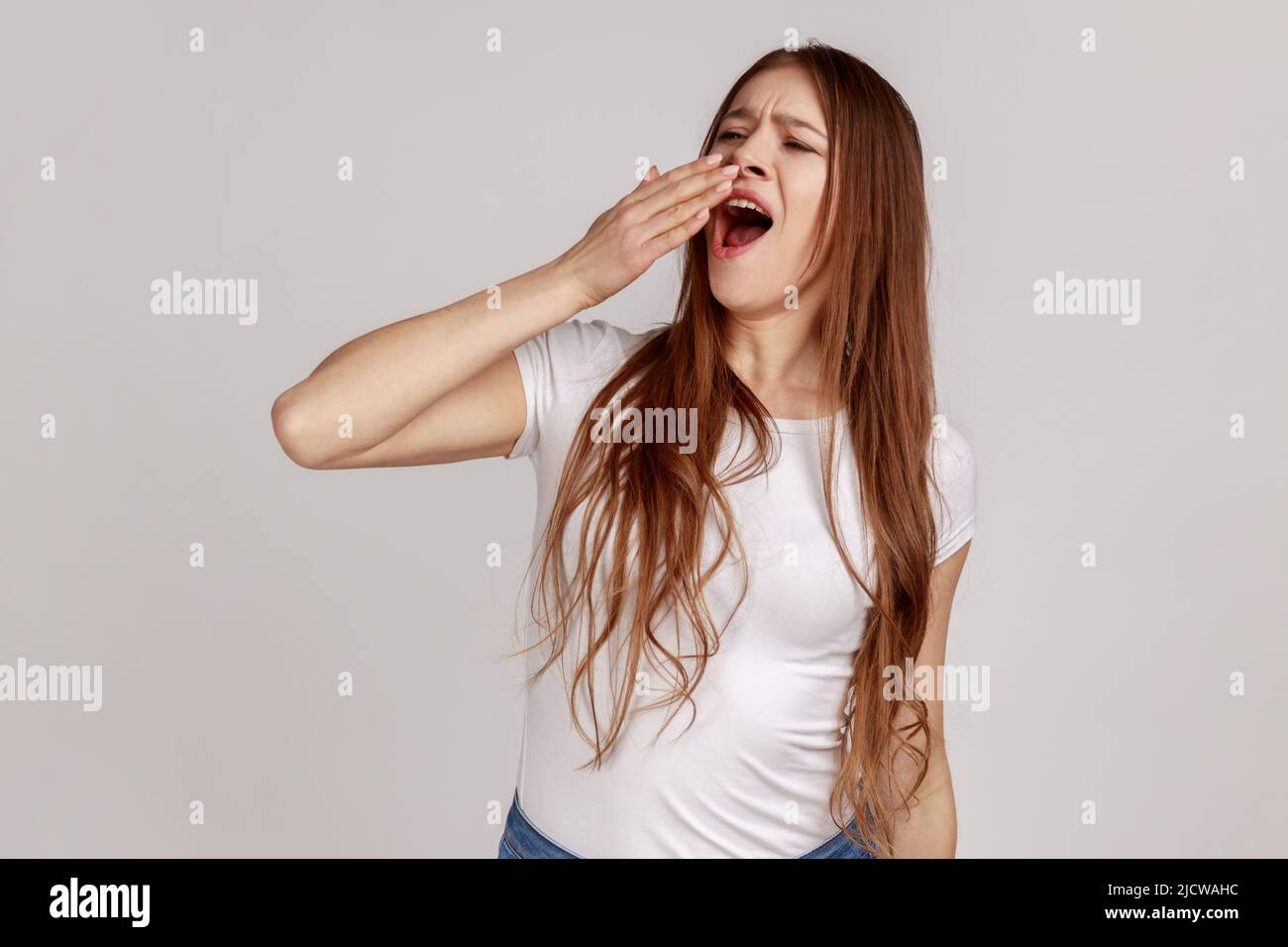 Porträt einer attraktiven müde, schlafenden Frau, die gähnt, den Mund mit der Hand schließt, erschöpft von Schlaflosigkeit, Mangel an Energie, mit weißem T-Shirt. Innenaufnahme des Studios isoliert auf grauem Hintergrund. Stockfoto