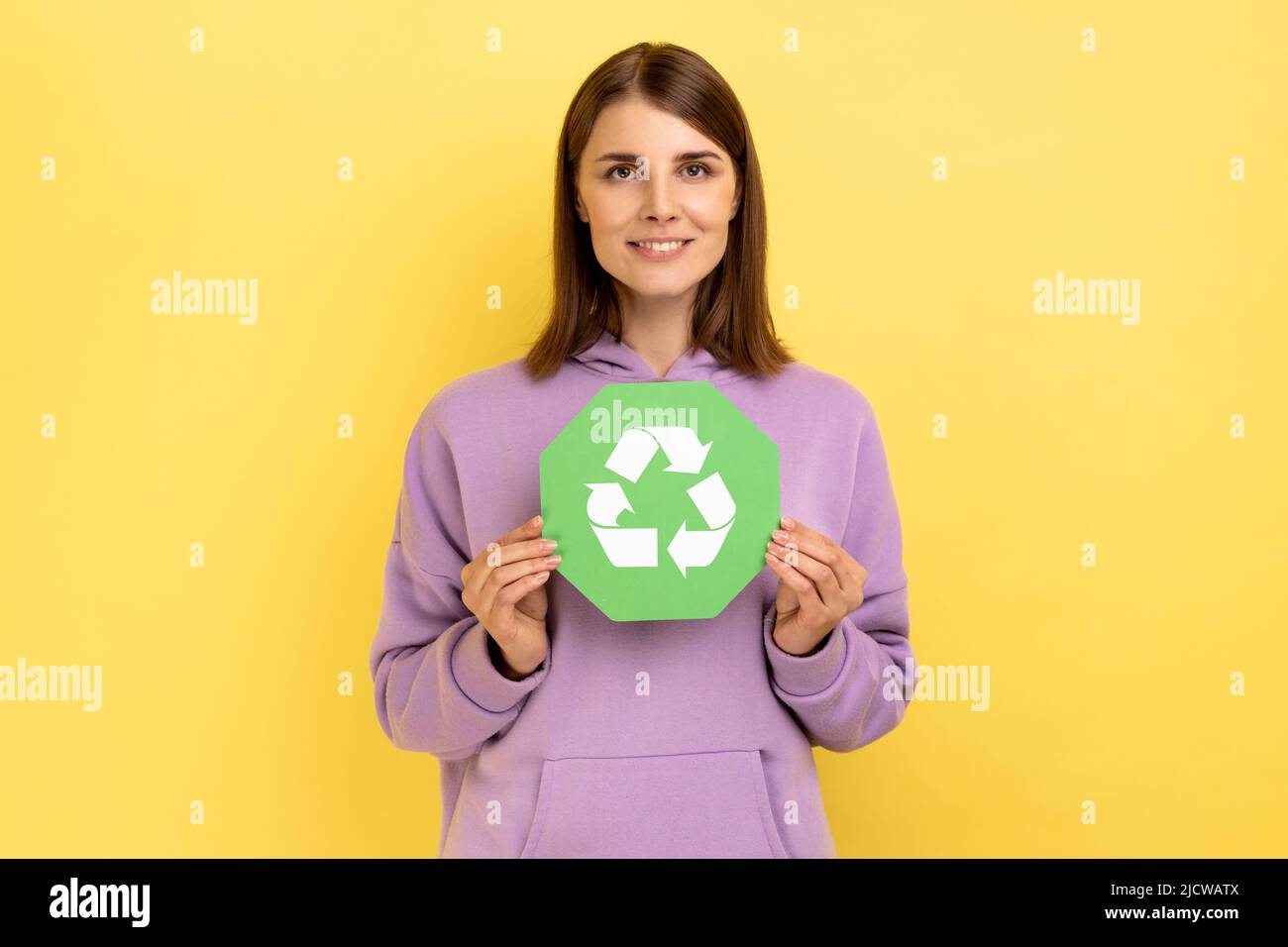 Lächelnde Frau mit dunklem Haar in den Händen halten grünes Recycling singen, Ökologie Konzept, Blick auf die Kamera, trägt lila Hoodie. Innenaufnahme des Studios isoliert auf gelbem Hintergrund. Stockfoto