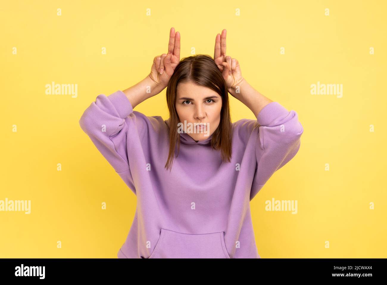 Porträt einer aggressiven Frau, die die Finger über den Kopf hält und Hörner zeigt, arrogant und stur, angriffbereit, mit purpurfarbenem Hoodie. Innenaufnahme des Studios isoliert auf gelbem Hintergrund. Stockfoto