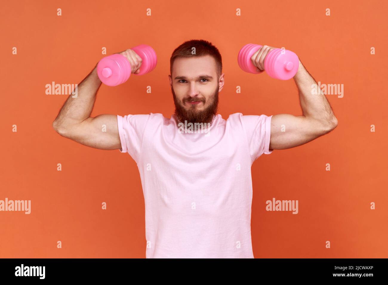 Porträt eines starken, athletisch aussehenden jungen bärtigen Mannes mit angehobenen Hanteln in erhobenen Armen, gesunder Lebensstil und rosa T-Shirt. Innenaufnahme des Studios isoliert auf orangefarbenem Hintergrund. Stockfoto