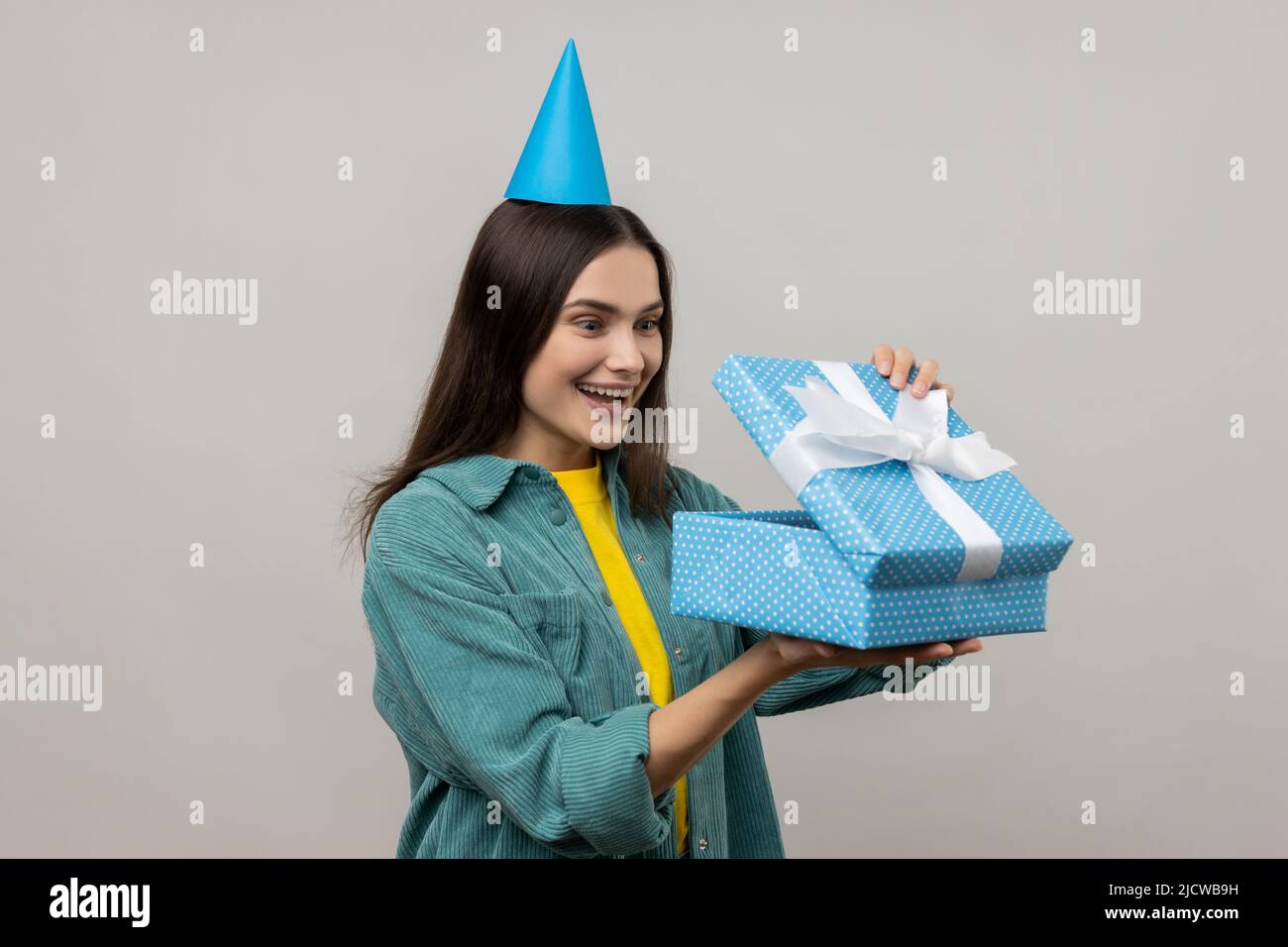 Porträt einer überraschten Frau mit Party-Kegel-Hut Blick in Geschenkbox, Eröffnung Geschenk und guckt nach innen mit glücklichen Lächeln, tragen lässigen Stil Jacke. Innenaufnahme des Studios isoliert auf grauem Hintergrund. Stockfoto