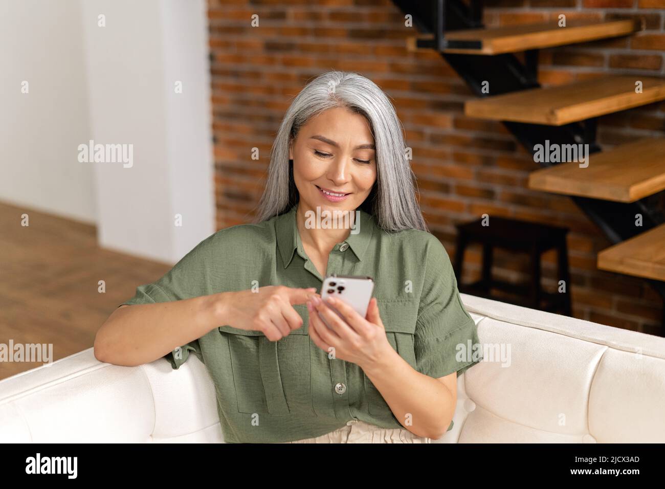 Ruhige ältere Dame, die in entspannter Pose auf der bequemen Couch mit einem Smartphone sitzt, verbringt die Frau ihre Freizeit damit, in den Neuigkeiten zu blättern und im Internet zu surfen Stockfoto