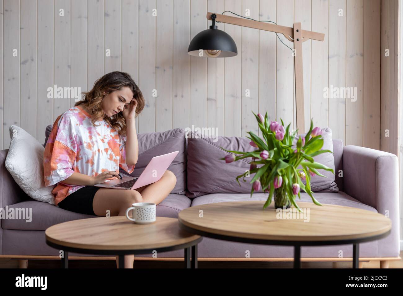 Junge charmante braunhaarige Frau mit langen Haaren in bequemer Heimkleidung arbeitet aufmerksam zu Hause im Internet auf einem gemütlichen Sofa sitzend. Modern Stockfoto