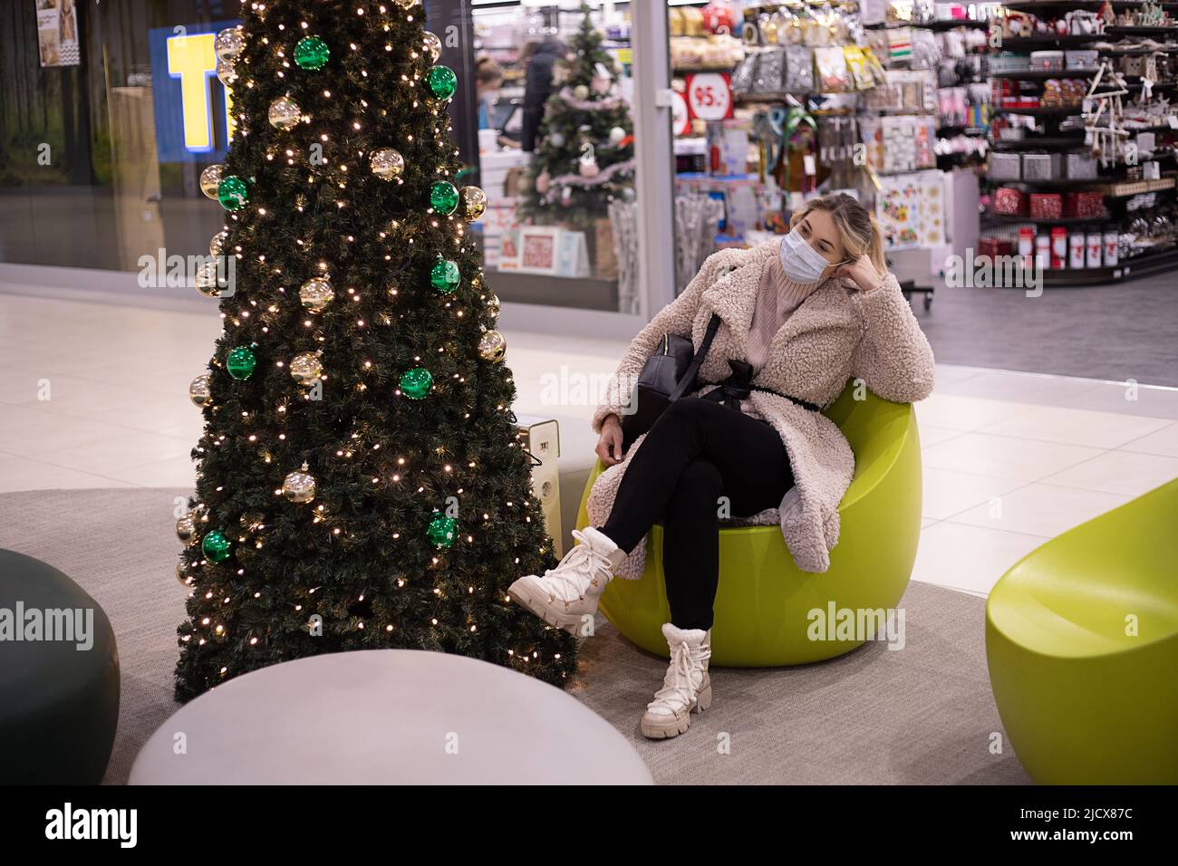 Hübsche Kundin in Maske, kuschelig weißer Wintermantel und hohe Stiefel sitzt entspannt im Sessel im Einkaufszentrum in der Nähe des Weihnachtsbaums. Einkaufszentrum Stockfoto