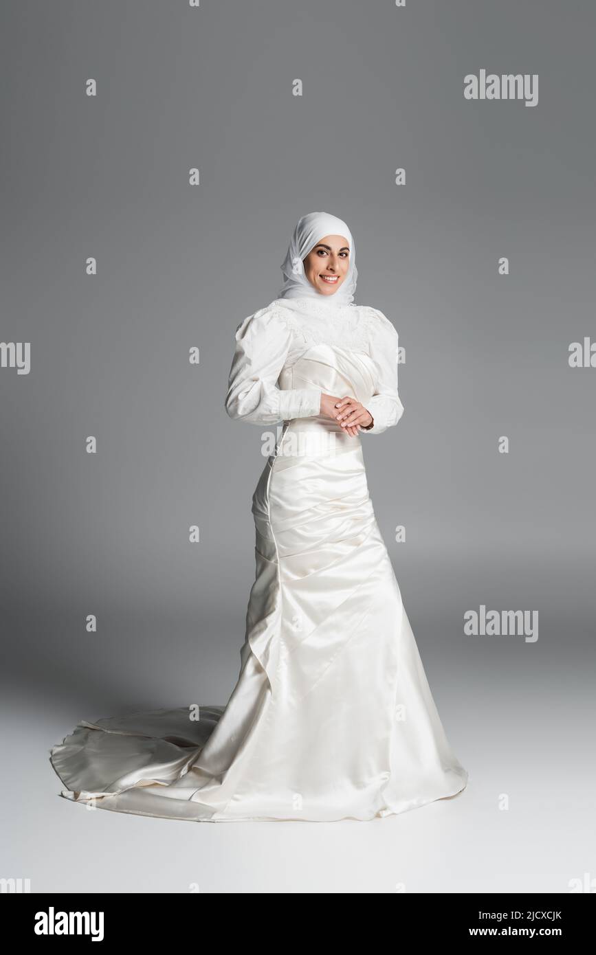 Die ganze Länge der glücklichen muslimischen Braut in Hochzeitskleid und  Hijab stehen auf dunkelgrau Stockfotografie - Alamy