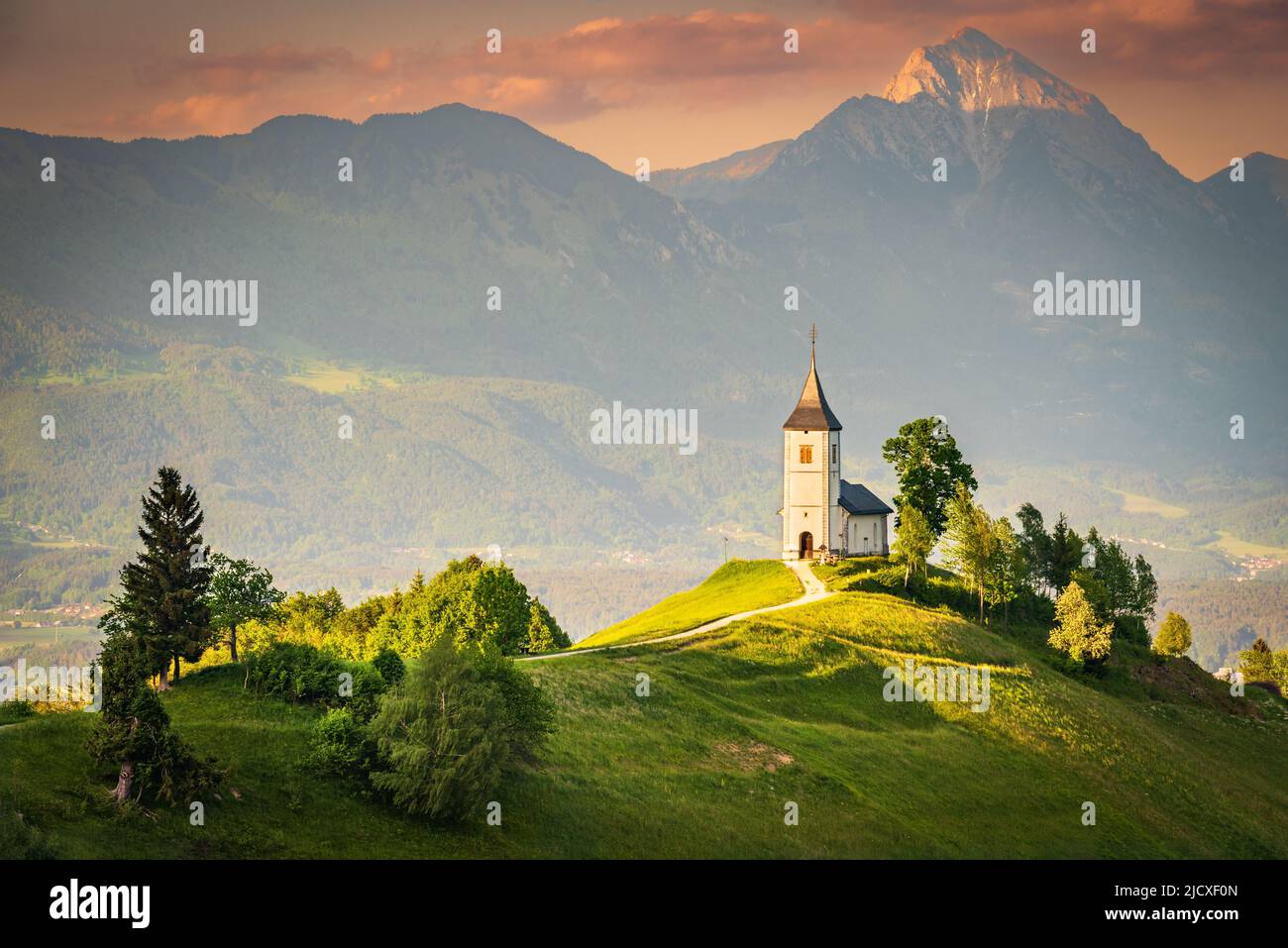 Jamnik, Slowenien - Luftaufnahme der wunderschönen Kirche auf dem Hügel, herrlicher goldener Sonnenuntergang über den Kamnik-Savinja Alpen, Skofja Loka Stockfoto