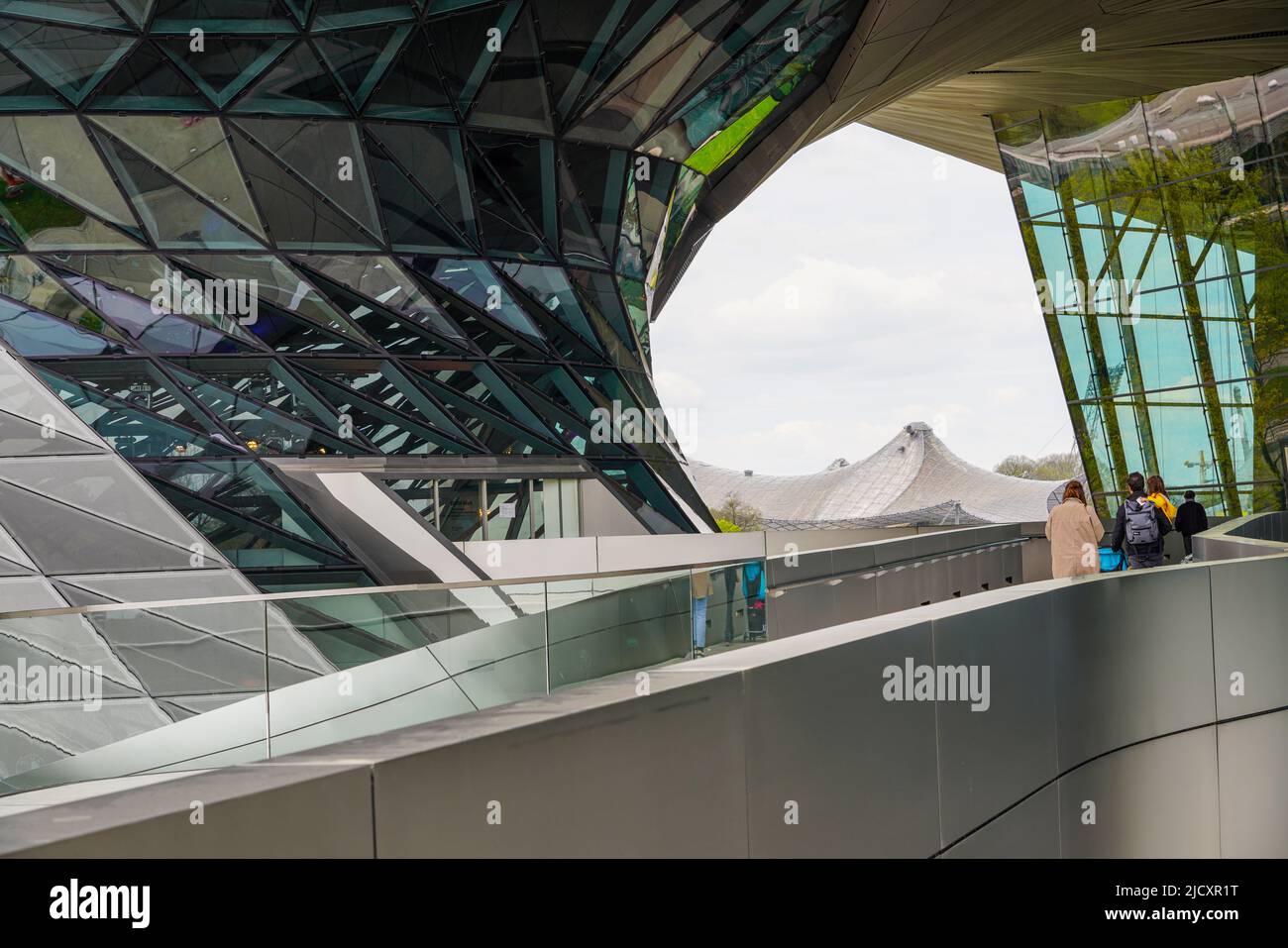 Ein Paar mit Kind tritt in die BMW Welt ein. Im Hintergrund die Dächer der Sportanlagen des Olympischen Zentrums, 19.4.22 Stockfoto
