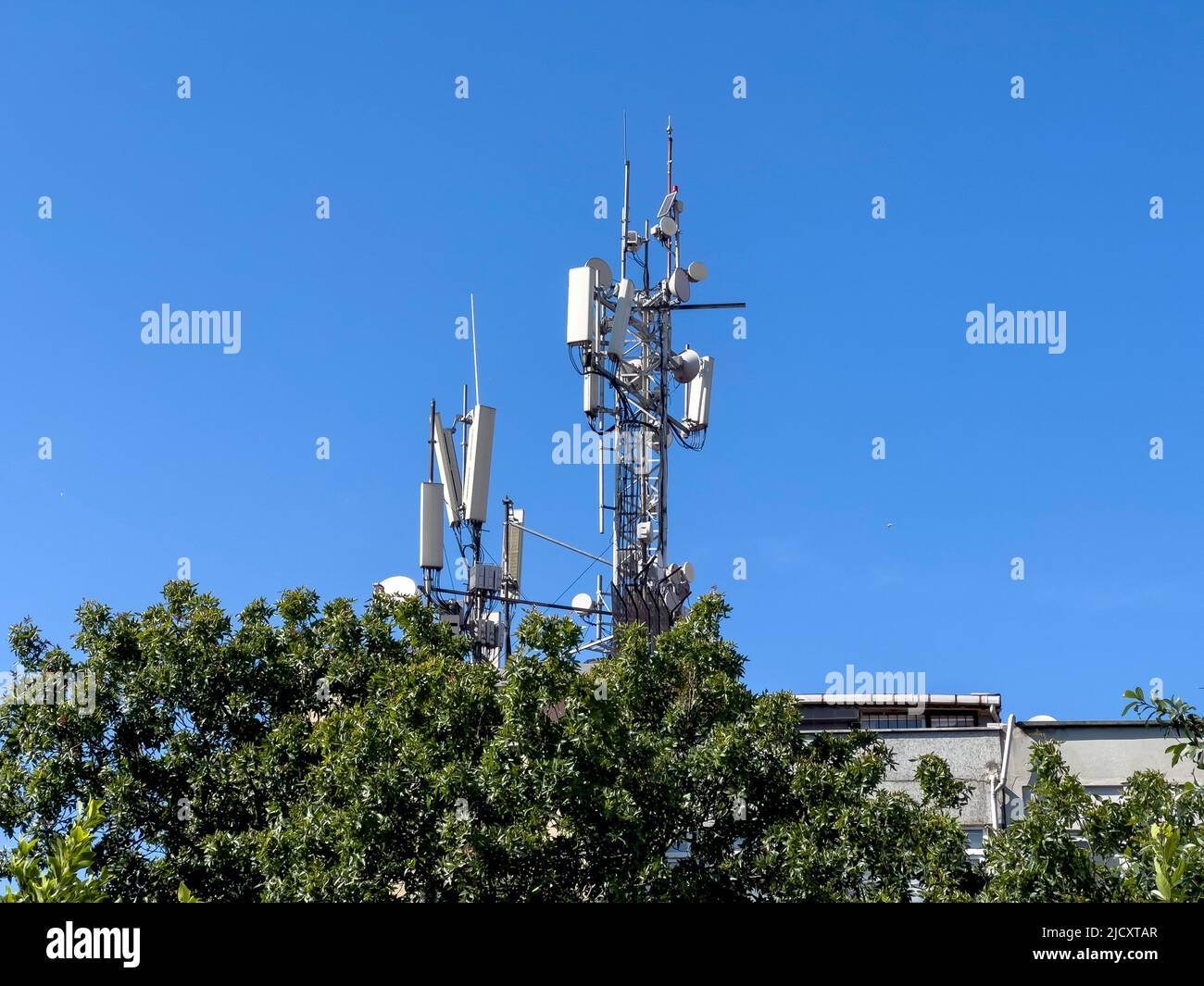 GSM-Antennenturm, Kopierplatz. Telekommunikationstechnologie für 5G, 4G, 3G. Mobiltelefon, TV-Antennen, Dach hinter grünem Baum. Sonniger blauer Himmel Stockfoto