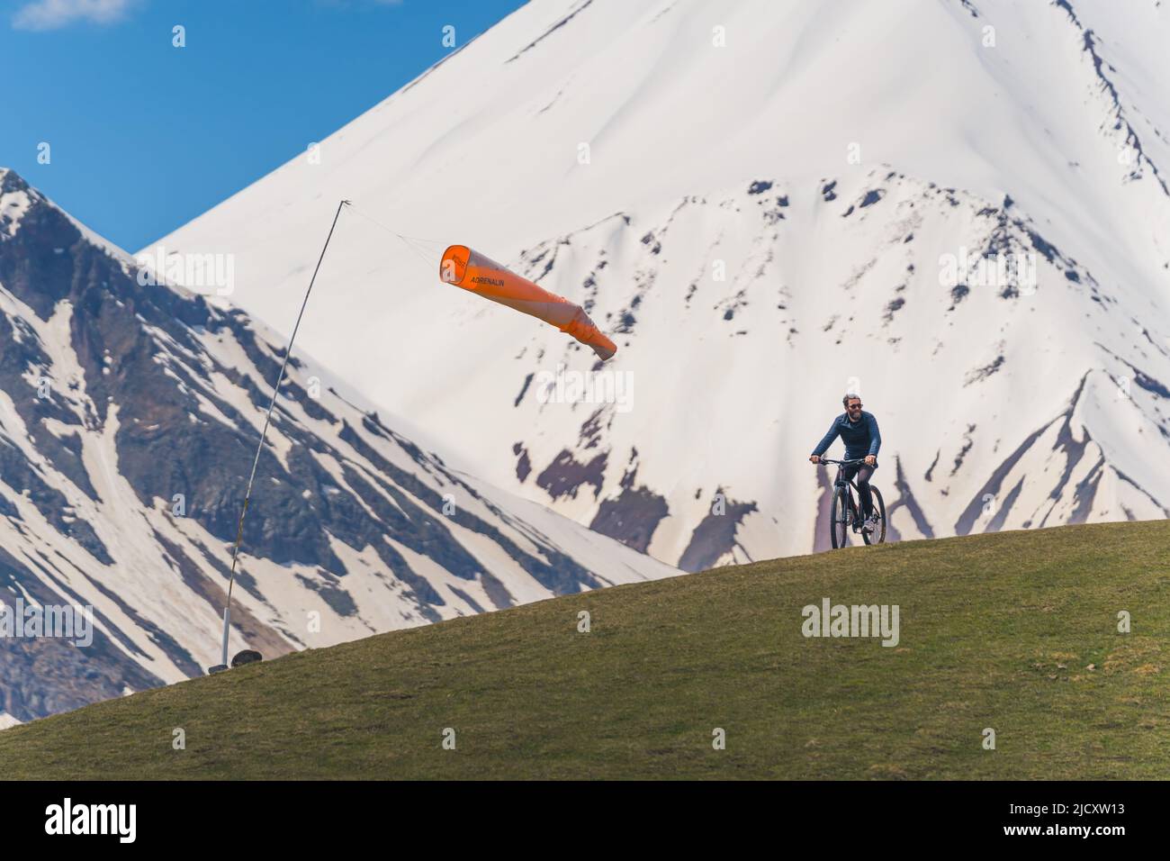 18.05.2022. Gudauri, Georgien. Junger Mann, der an einem sonnigen Sommertag auf einer Bergwiese Fahrrad fährt, schneebedeckte Kaukasus-Berge im Hintergrund. Hochwertige Fotos Stockfoto