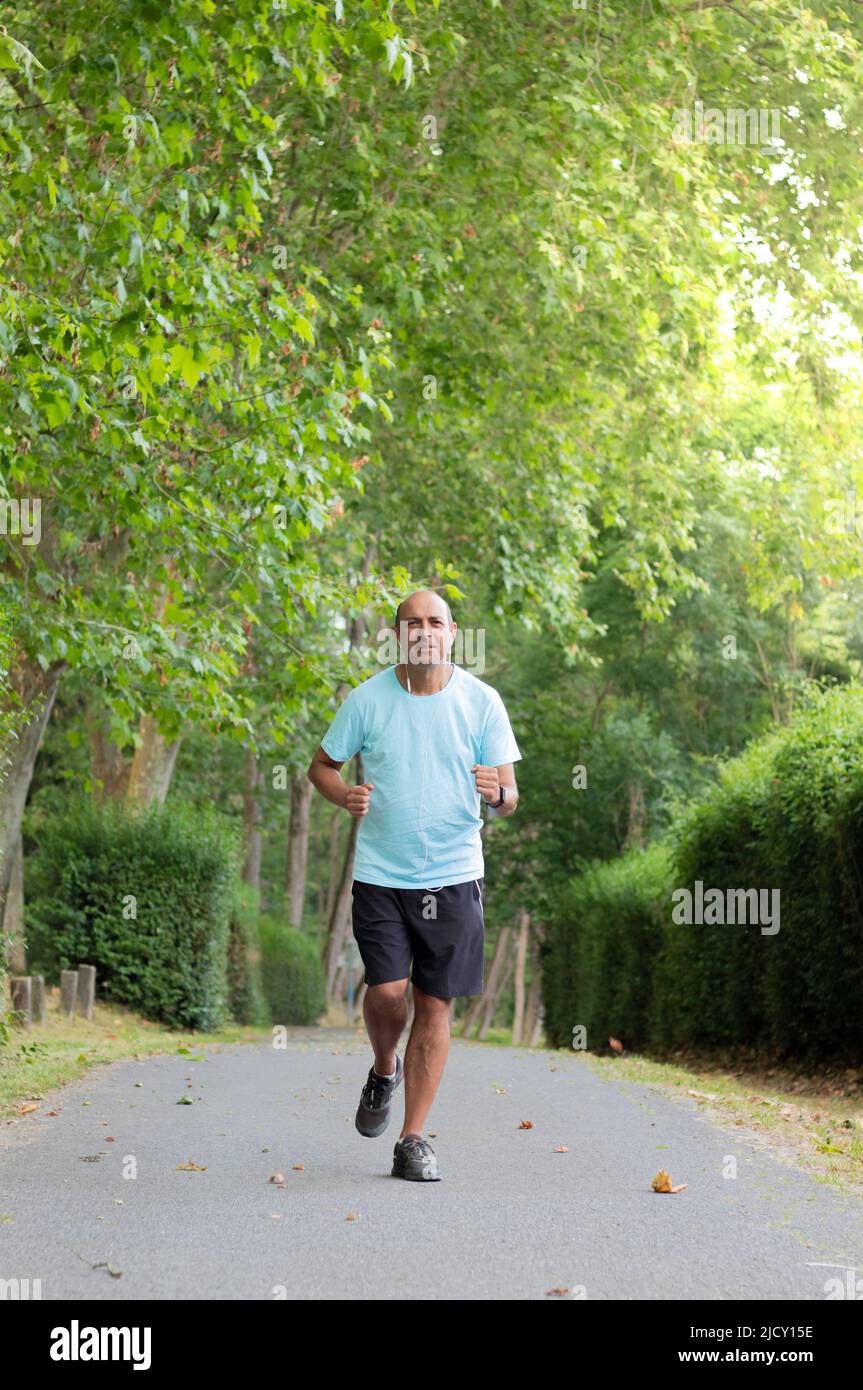 Reifer, haarloser Mann in Sportbekleidung joggt auf einem Betonweg zwischen den Büschen und Bäumen der Natur Stockfoto