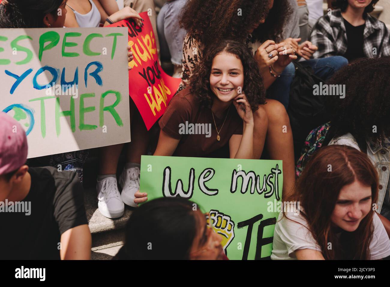 Ein fröhliches Teenager-Mädchen lächelt an der Kamera und hält ein Banner, während es mit einer Gruppe von Klimaaktivisten sitzt. Diverse Demonstranten, die für Klam aufschlagen Stockfoto