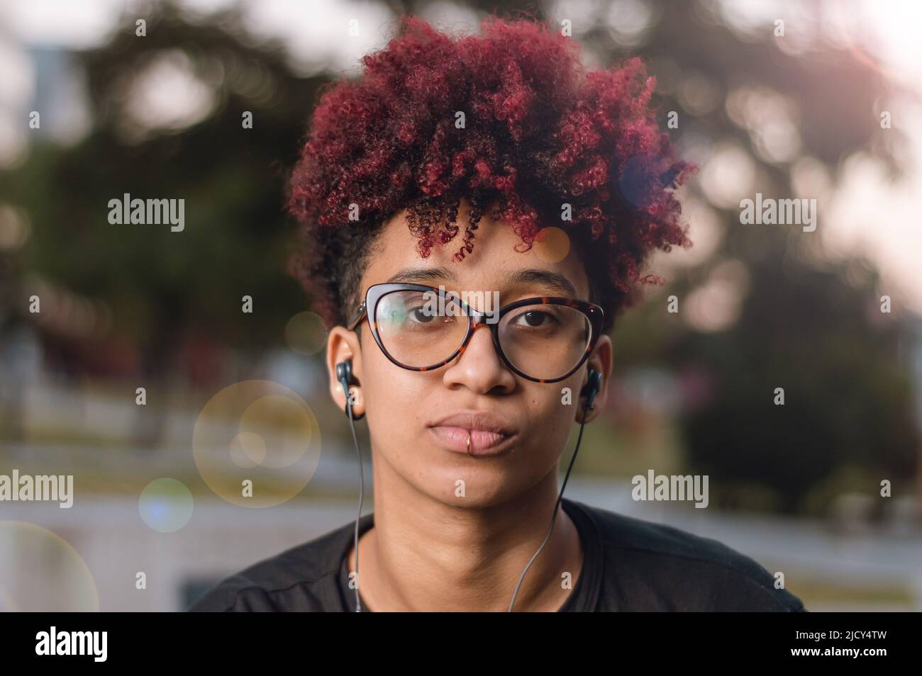 Nahaufnahme Porträt im Freien schöne junge lateinamerikanische Brünette Frau mit gefärbtem roten Afro, Brille und Kopfhörer, ernsthaft Blick auf die Kamera Stockfoto