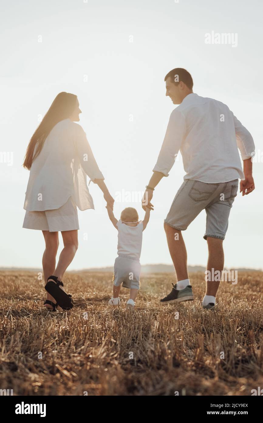 Rückansicht von Mama und Papa, die ihrem Kind beim ersten Schritt helfen, junge Familie beim Sonnenuntergang auf dem Feld Stockfoto