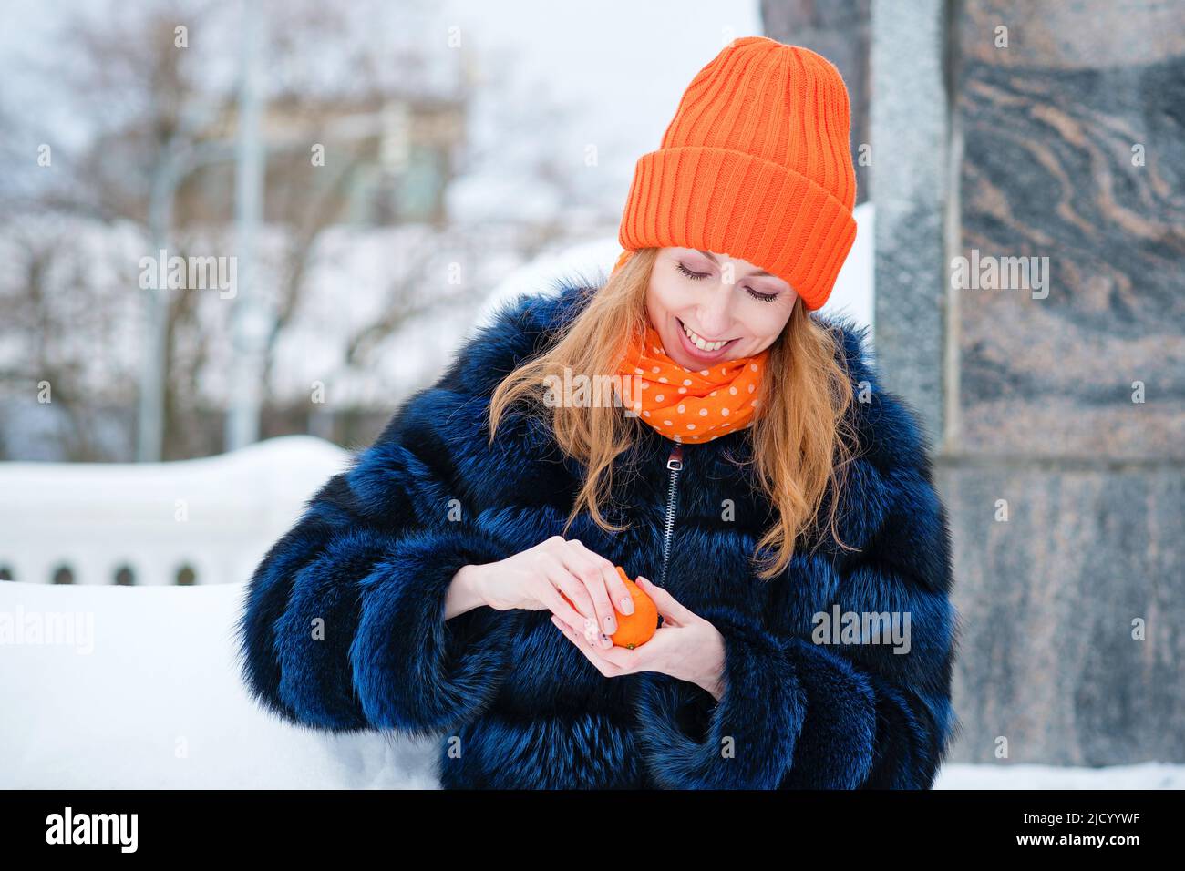 Attraktive Mädchen zu Fuß im Winter Park. Junge Frau im stilvollen warmen Outfit und Orange Beanie Hut Peeling Tangerine und Blick auf Obst im Freien. Stockfoto