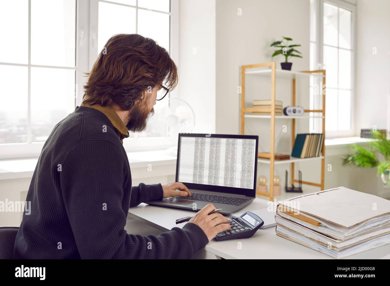 Männlicher Buchhalter, der im Büro sitzt, mit Tabellenkalkulationen auf dem Laptop arbeitet und Rechner benutzt Stockfoto