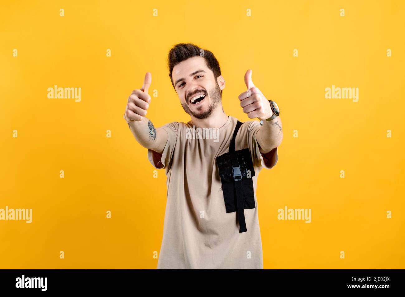 Porträt eines Mannes, der beige T-Shirts trägt und isoliert auf gelbem Hintergrund posiert und Handgesten für großartige Leistung tut. Zeigen Sie, dass beide Hände Daumen nach oben zeigen Stockfoto