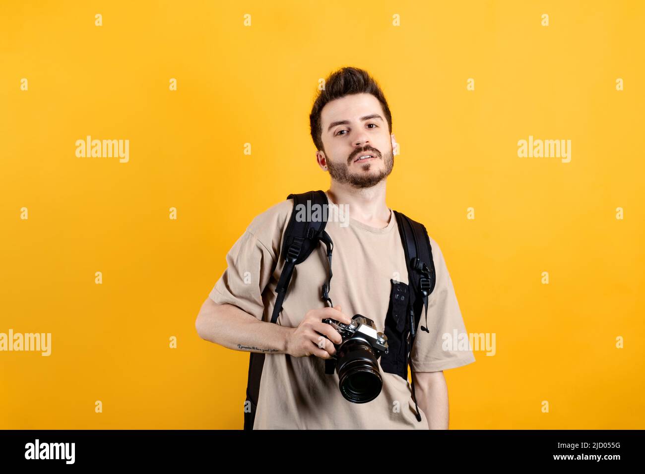 Junger Mann mit beigefarbenem T-Shirt, der isoliert auf gelbem Hintergrund posiert und mit seiner dslr-Kamera und seiner professionellen Fotoausrüstung posiert. Stockfoto