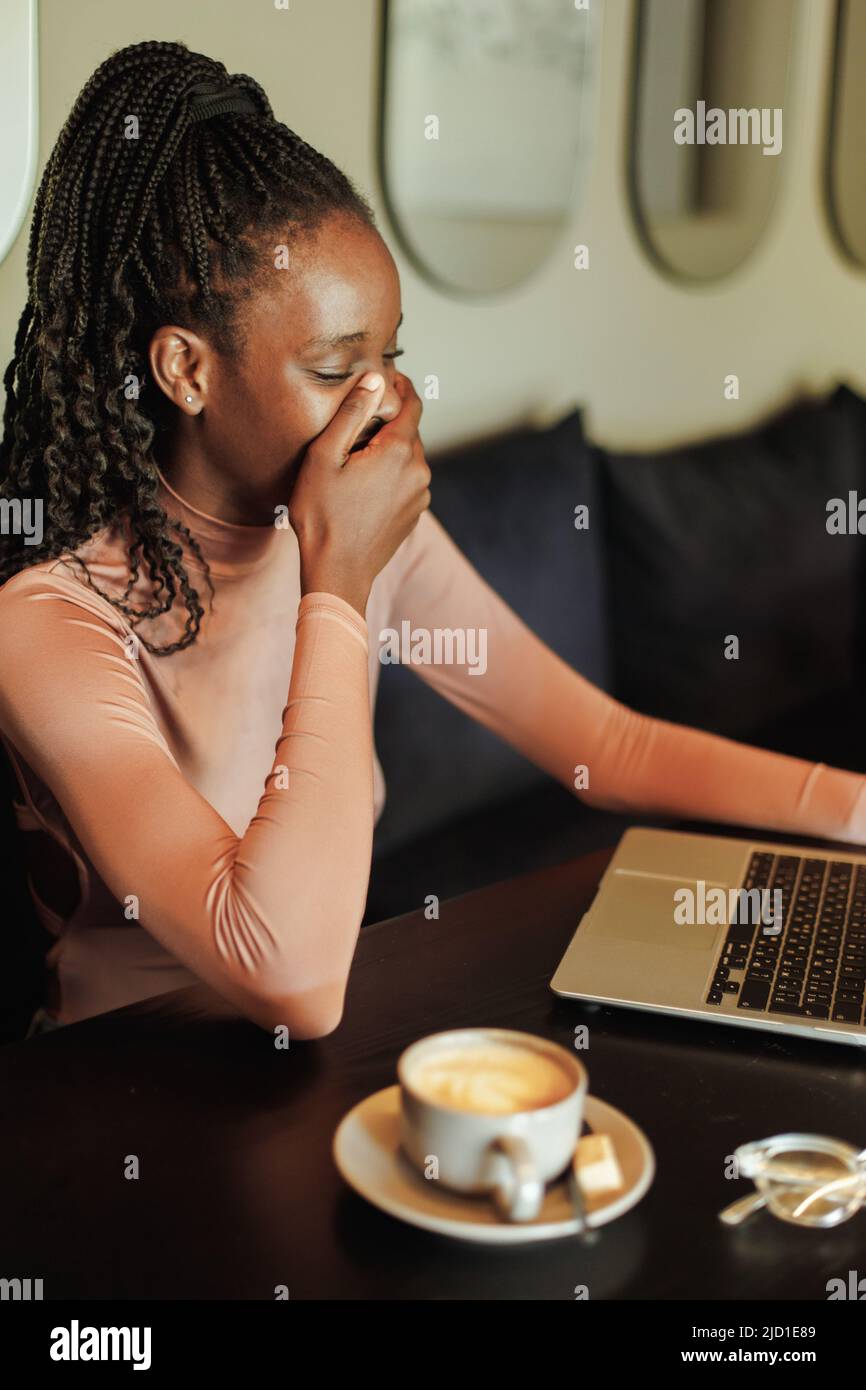 Lachende tausendjährige afro-amerikanische Bloggerin, den Mund mit der Hand bedeckend, Kaffee trinken, Webinar auf Laptop im Café ansehen Stockfoto