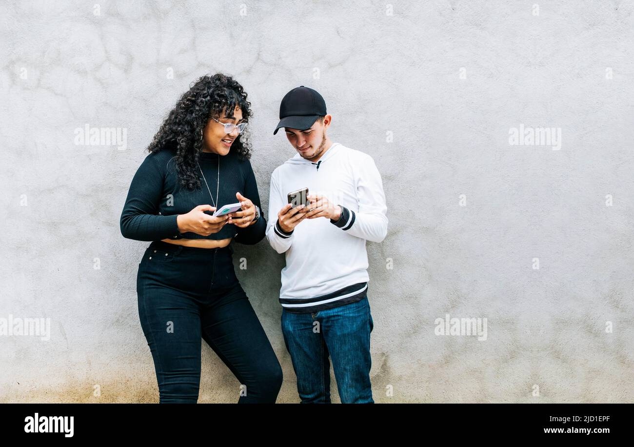 Zwei Teenager-Freunde, die ihre Handys checken und lächeln, zwei Teenager, die zusammen ihre Handys checken, Guy und Mädchen, die sich an einer Wand lehnen Stockfoto