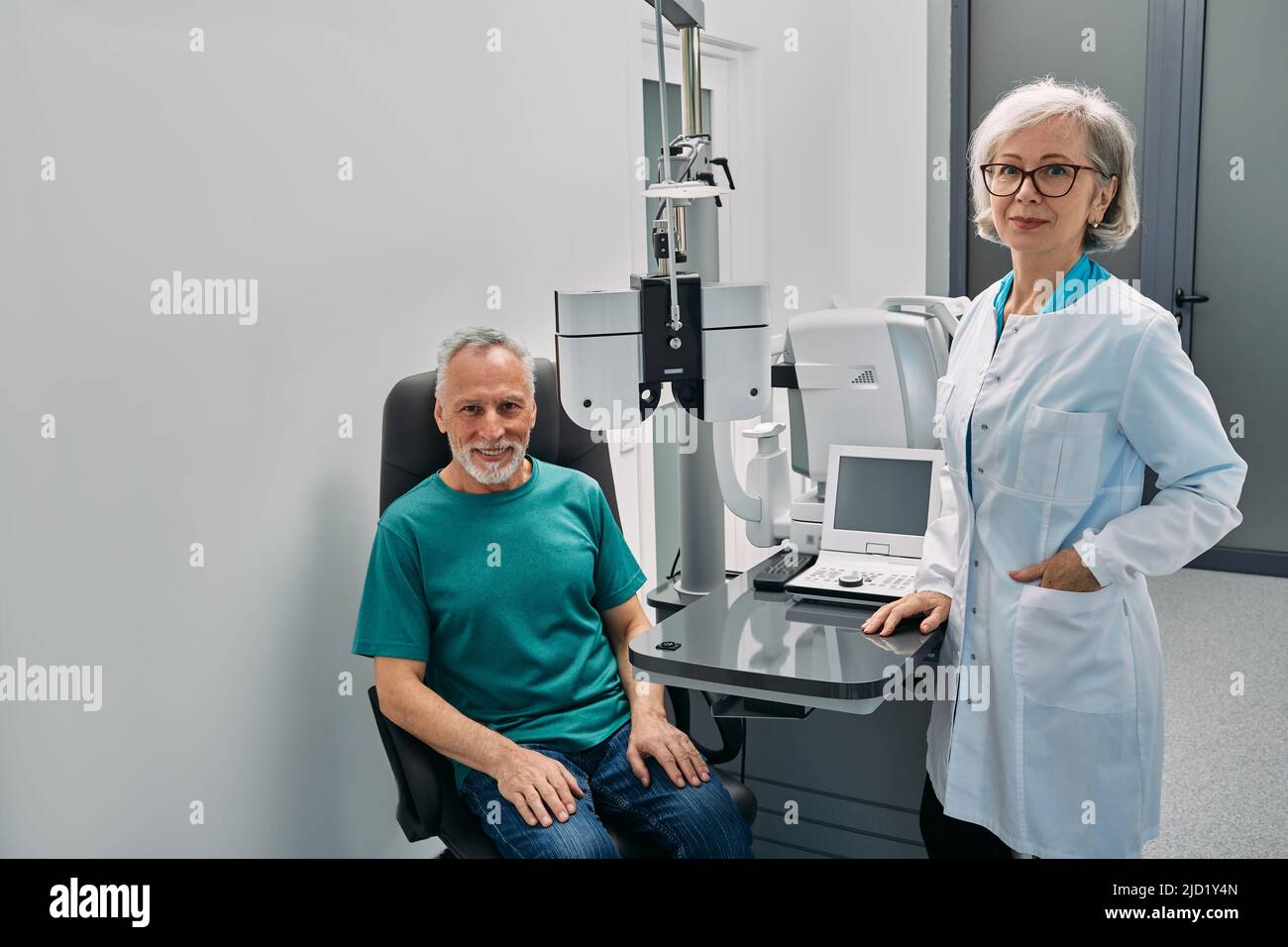 Positive Optometristin mit ihrem älteren männlichen Patienten, der während einer ophthalmologischen Konsultation in der Nähe von ophthalmischen Geräten freundlich auf die Kamera schaut Stockfoto
