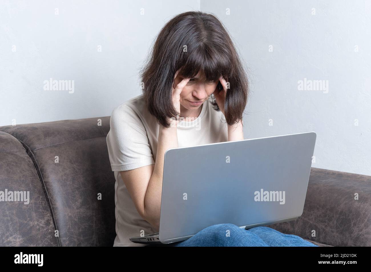 Frustrierte, traurige Frau mittleren Alters fühlt sich müde, massiert ihre Schläfen, sorgt sich um ein Problem, während sie mit einem Laptop auf einem Sofa sitzt. Arbeitsüberstunden Stockfoto