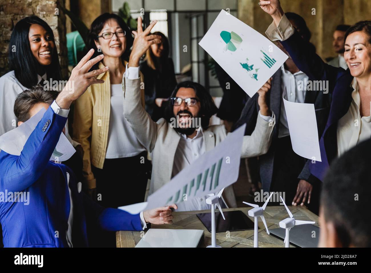 Multiethnische Geschäftsleute feiern den Erfolg eines Projekts im Bereich erneuerbare Energien und werfen gemeinsam Dokumente ins Büro - Windmühlen im Fokus Stockfoto