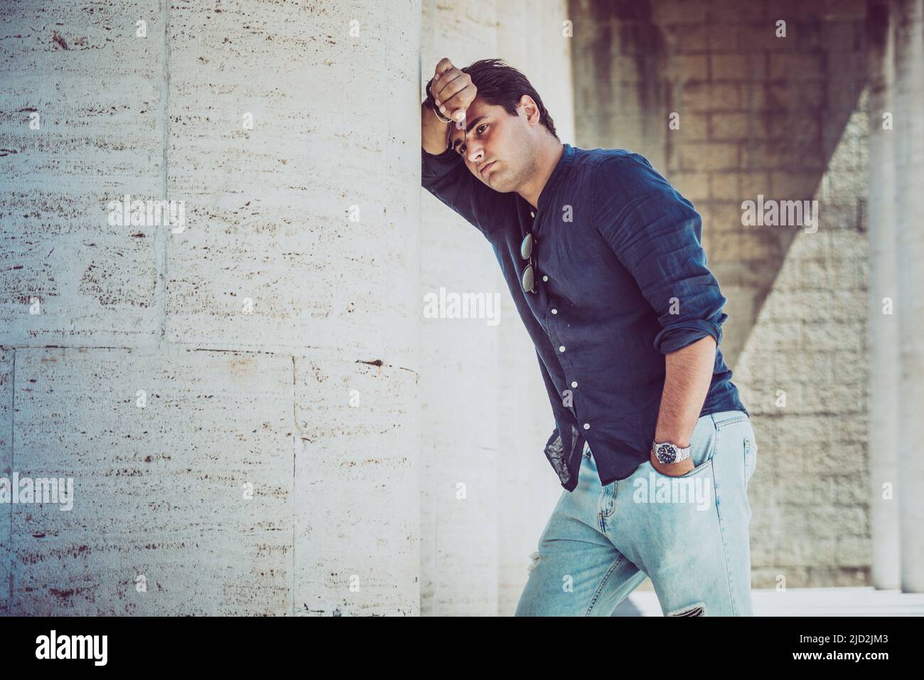 Junger depressiver und müder Mann im Freien, der mit einem Arm an einer weißen Wand gelehnt ist. Lässige Kleidung. Stockfoto