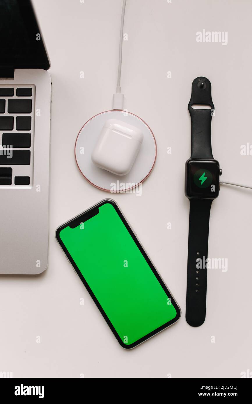 Arbeitsplatz mit Laptop auf Tisch und Geräte. Kabelloses Aufladen von Smart Watch und kabellosen Kopfhörern auf weißem Tisch. Smartphone mit grünem Bildschirm Stockfoto