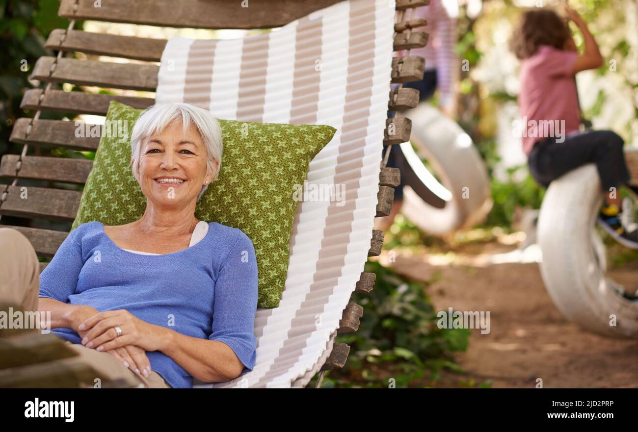 Auch die Großeltern müssen sich wieder aufladen. Porträt einer älteren Frau, die in einer Hängematte im Garten liegt, während ihre Enkel im Hintergrund spielen. Stockfoto