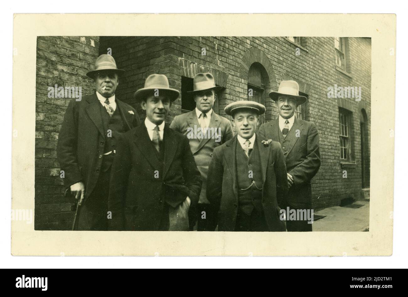 Ursprüngliche Postkarte aus den frühen 1920er Jahren einer Gruppe von Männern der Arbeiterklasse, die für ein Foto in einer städtischen Straße, in einer armen Nachbarschaft posieren, Fedora- oder Trilby-Hüte tragen und ein Mann eine flache Mütze, Peaky-Blinders/Gangster-Lookalikes, möglicherweise eine Hochzeitsfeier trägt, Bleibt zurück, während der Mann vorne eine Knopflochblume auf dem Revers trägt. Ca. 1925. GROSSBRITANNIEN Stockfoto