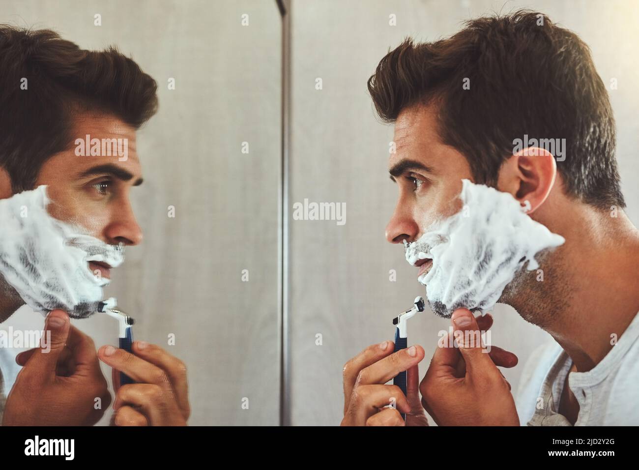 Schön und nahe gelegen. Eine kurze Aufnahme eines hübschen jungen Mannes, der sich den Bart rasieren lässt, während er zu Hause im Spiegel seine Spiegelung betrachtet. Stockfoto
