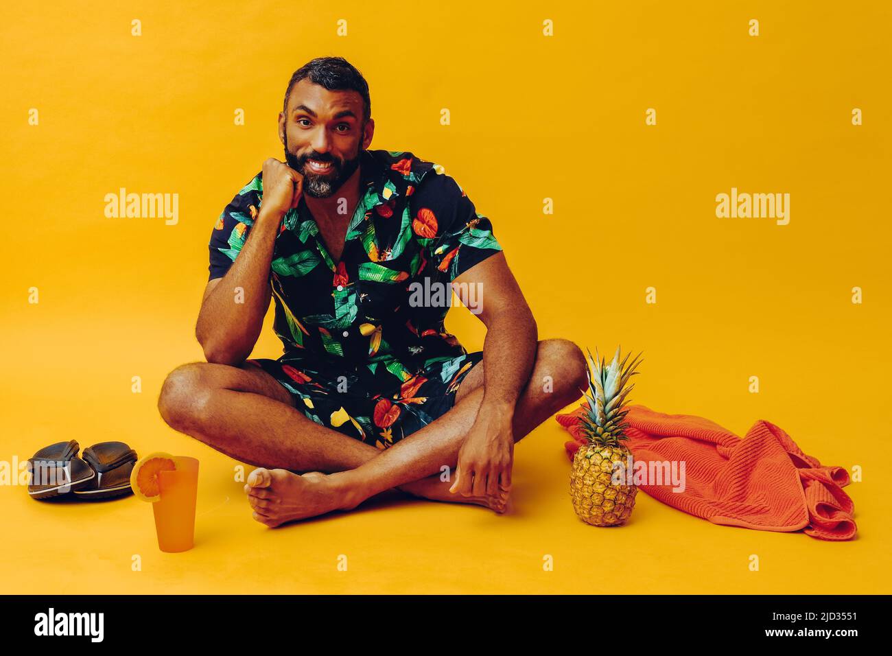 Schöner bärtiger afroamerikanischer Mann mit mittlerem Erwachsenen, der im Urlaub lächelt und mit einem orangen Handtuch, Orangensaft, Ananas und Sandalen im Studio sitzt Stockfoto