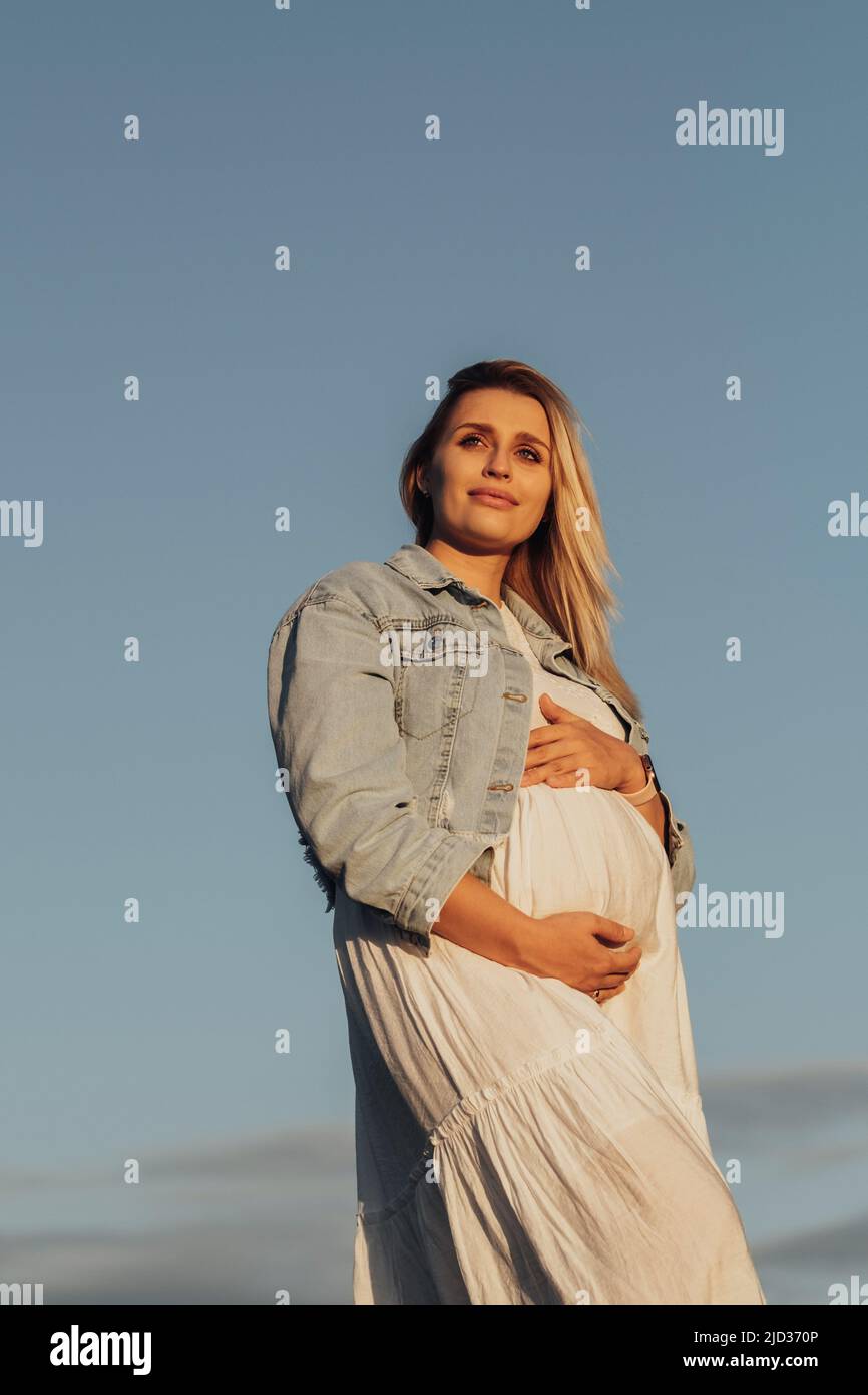 Porträt einer Schwangeren jungen kaukasischen Frau, die ihren Bauch auf dem Hintergrund des blauen Himmels hält Stockfoto