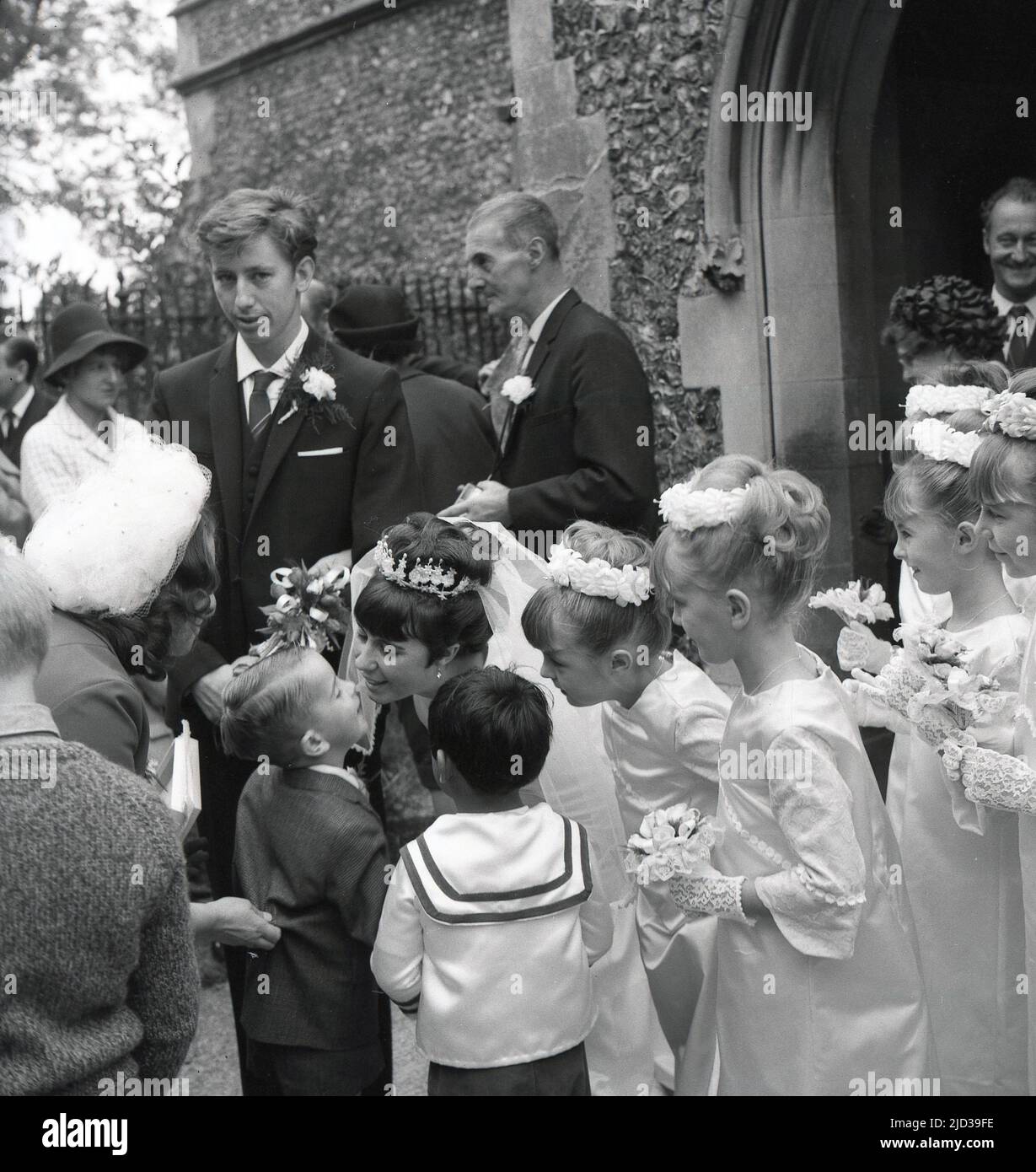1960s, historisch, vor einem Kircheneingang, eine gerade verheiratete Braut, umgeben von ihren jungen Brautjungfern, die einen Kuss von einem Page Boy, England, Großbritannien, bekommen. Stockfoto