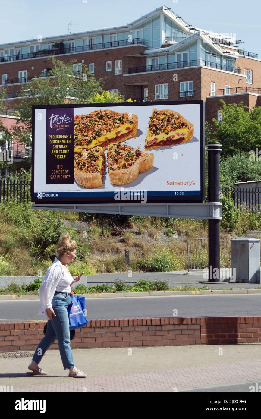 Eine Frau wartet, um die Straße unter einer digitalen Plakatwand zu überqueren, die sainsbury's Pflugmann's quiche in kingston upon thames, surrey, england, annonciert Stockfoto