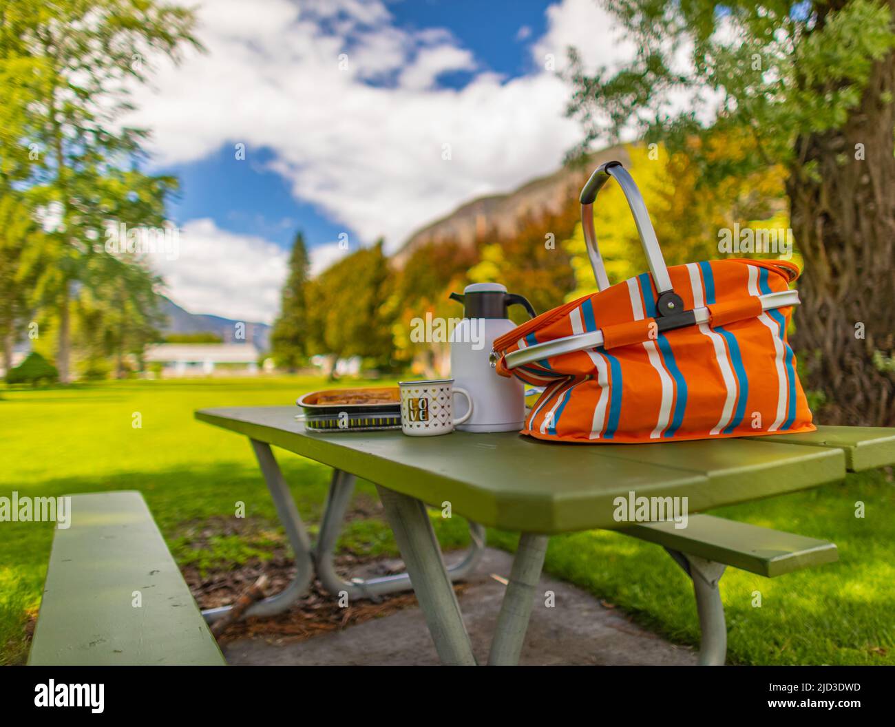 Picknick im Sommer. Holztisch mit Kisten und Körben mit Lebensmitteln für das Picknick im Freien in einem Park am blauen Himmel und Wolken im Hintergrund. Blick auf die Straße, Stockfoto
