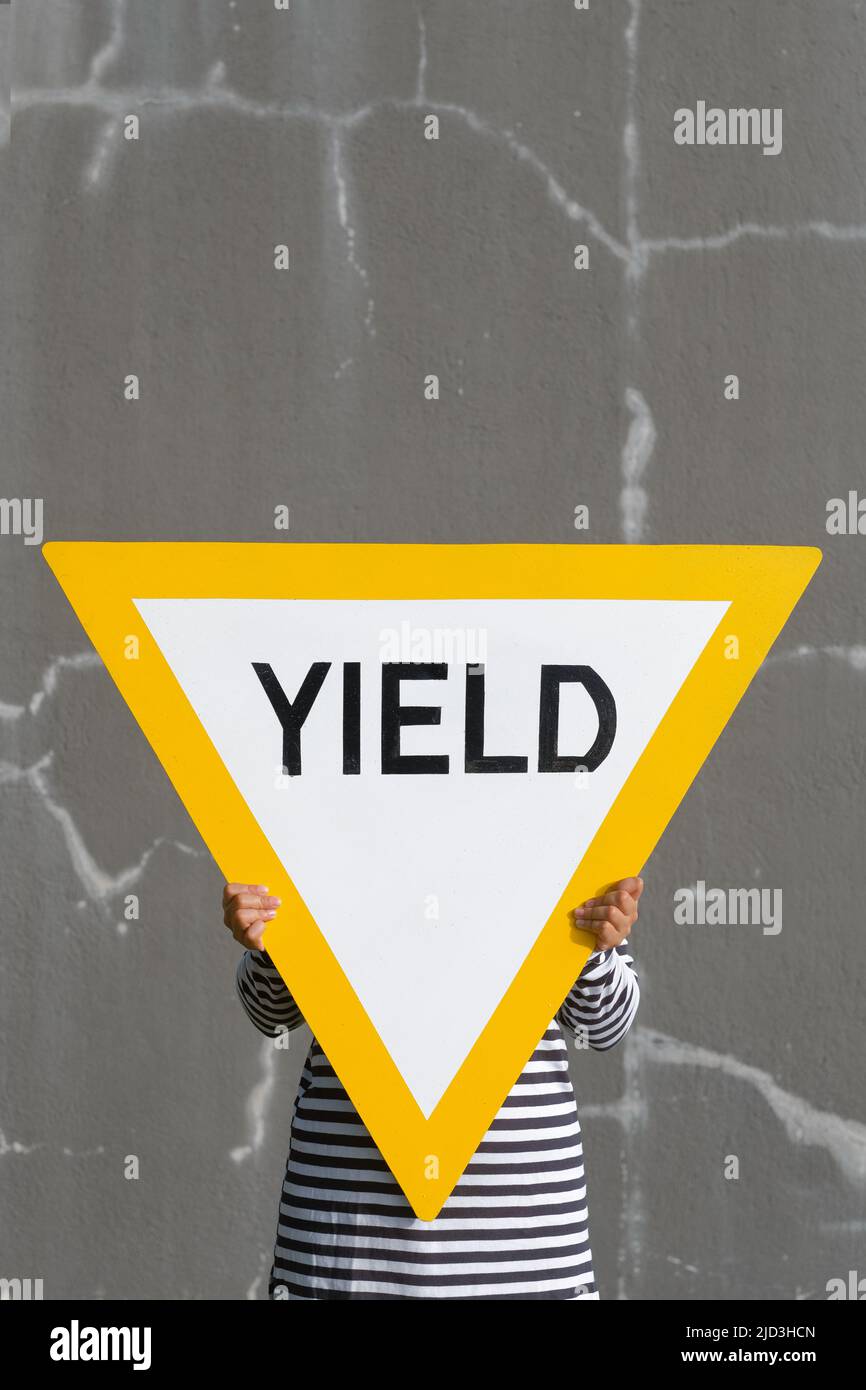 Person hält großes Rendite-Straßenschild. Kräftiges Gelb gegen rissige graue Wände. Symbol für abstrakte Konzepte: Give Way, Concede, Profit, Revenue, Harvest. Stockfoto