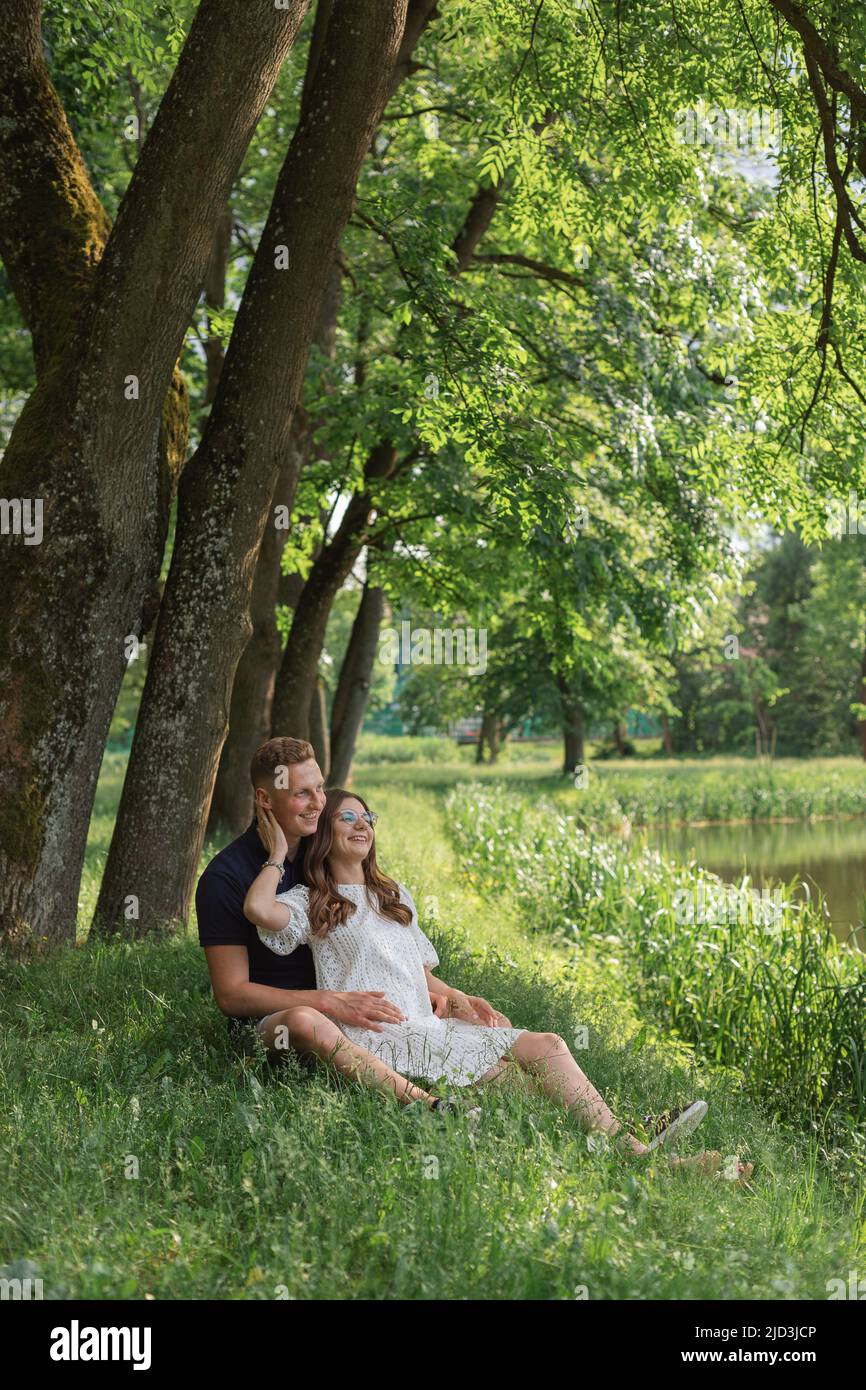 Niedliches junges Paar, das auf Gras unter dem Baum in der Nähe des Sees sitzt und sich gegenseitig anschaut. Junger Mann Frau genießen Sommertag. Stockfoto