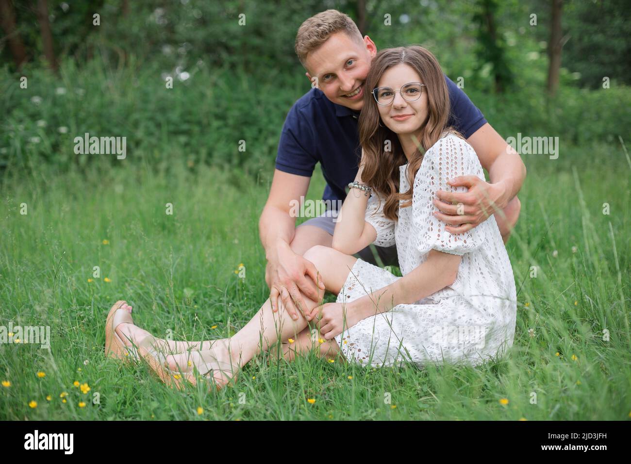 Liebevolles junges Paar, das im Park auf Gras sitzt und die Kamera anschaut. Junger Mann, Frau am Sommertag, Honigmond Stockfoto