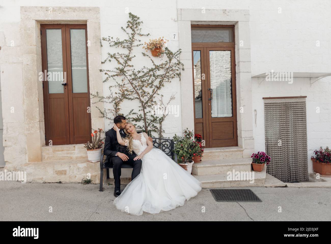 Portrait der Hochzeit, verliebtes Ehepaar, sitzend auf der Bank in der Nähe von Türen der Kirche. Pflanzen und Bäume an der Wand Stockfoto