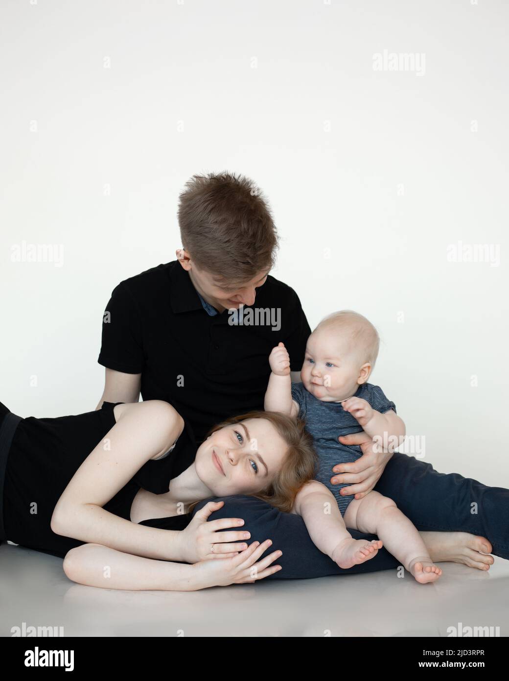 Junge Mama und Papa sitzen und umarmen mit Baby in dunkler Kleidung, lächeln und genießen auf weißem Hintergrund. Nette Beziehung zwischen Eltern und Kleinkind Stockfoto