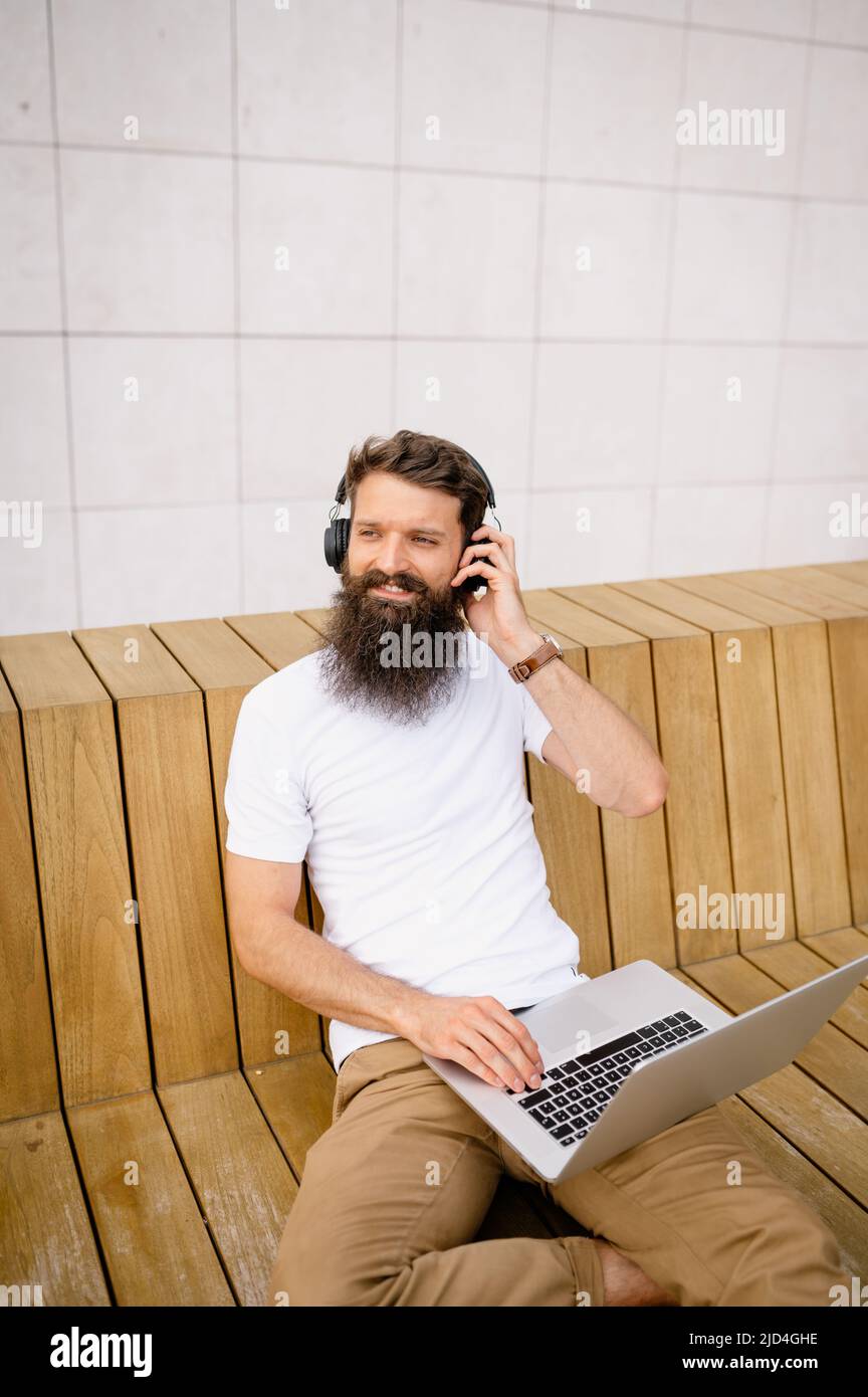 Lächelnder Mann mit mittlerem Erwachsenenalter, der auf der Bank sitzt und einen Laptop benutzt Stockfoto